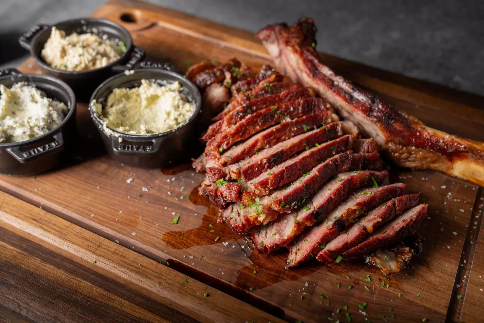 Sliced grilled steak on a wooden board with small bowls of condiments, including butter.