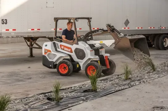 A man is driving a small bobcat loader  next to a semi truck.