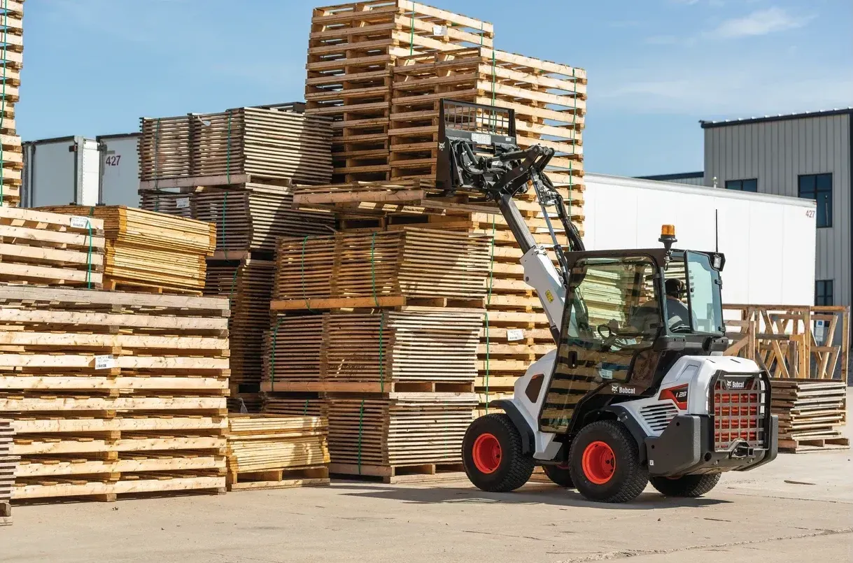 A man is driving a bobcat forklift in front of a pile of wooden pallets.