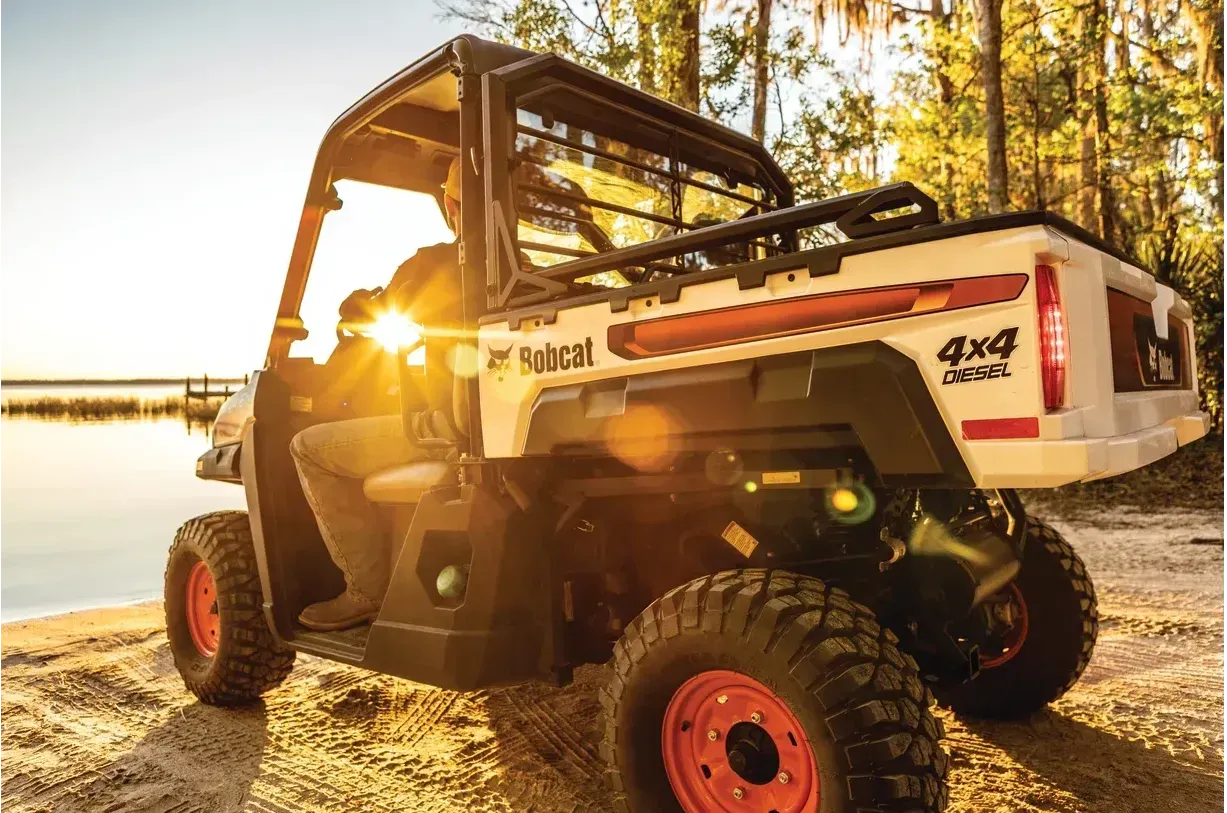 Un vehículo utilitario Bobcat está estacionado en la playa junto a un cuerpo de agua.