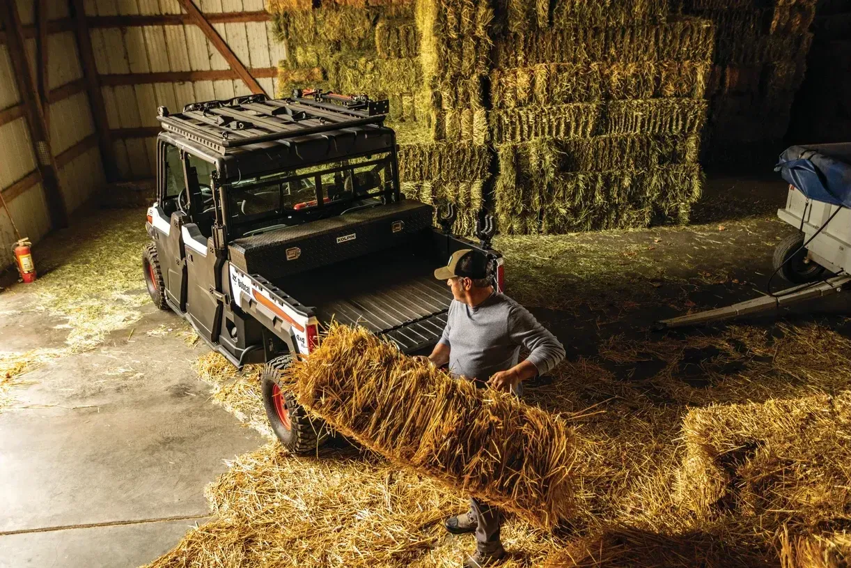 A man is loading hay into a bobcat side by side in a barn.