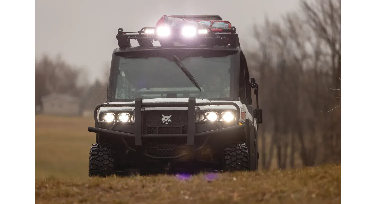 A bobcat side by side  with lights on top of it is driving down a dirt road.