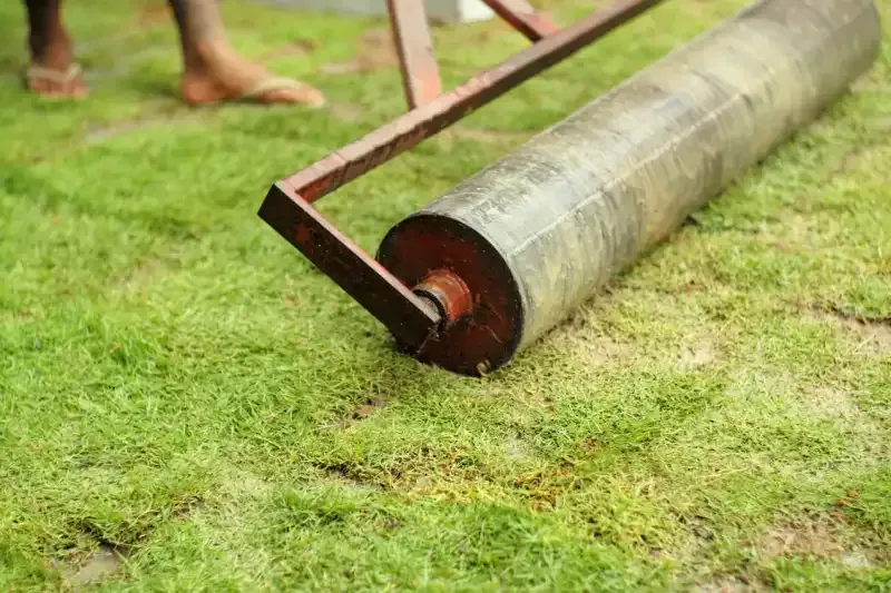 Person rolling a heavy, cylindrical lawn roller across green grass.
