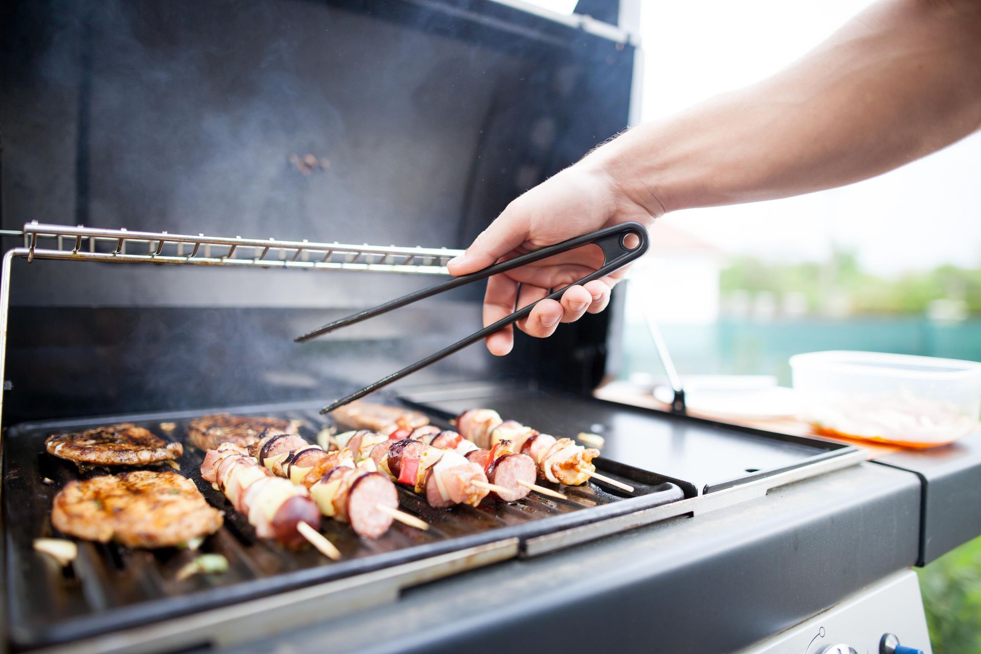Person grilling food on a barbecue; kebabs, meat, and mushrooms on the grill, tongs in use.
