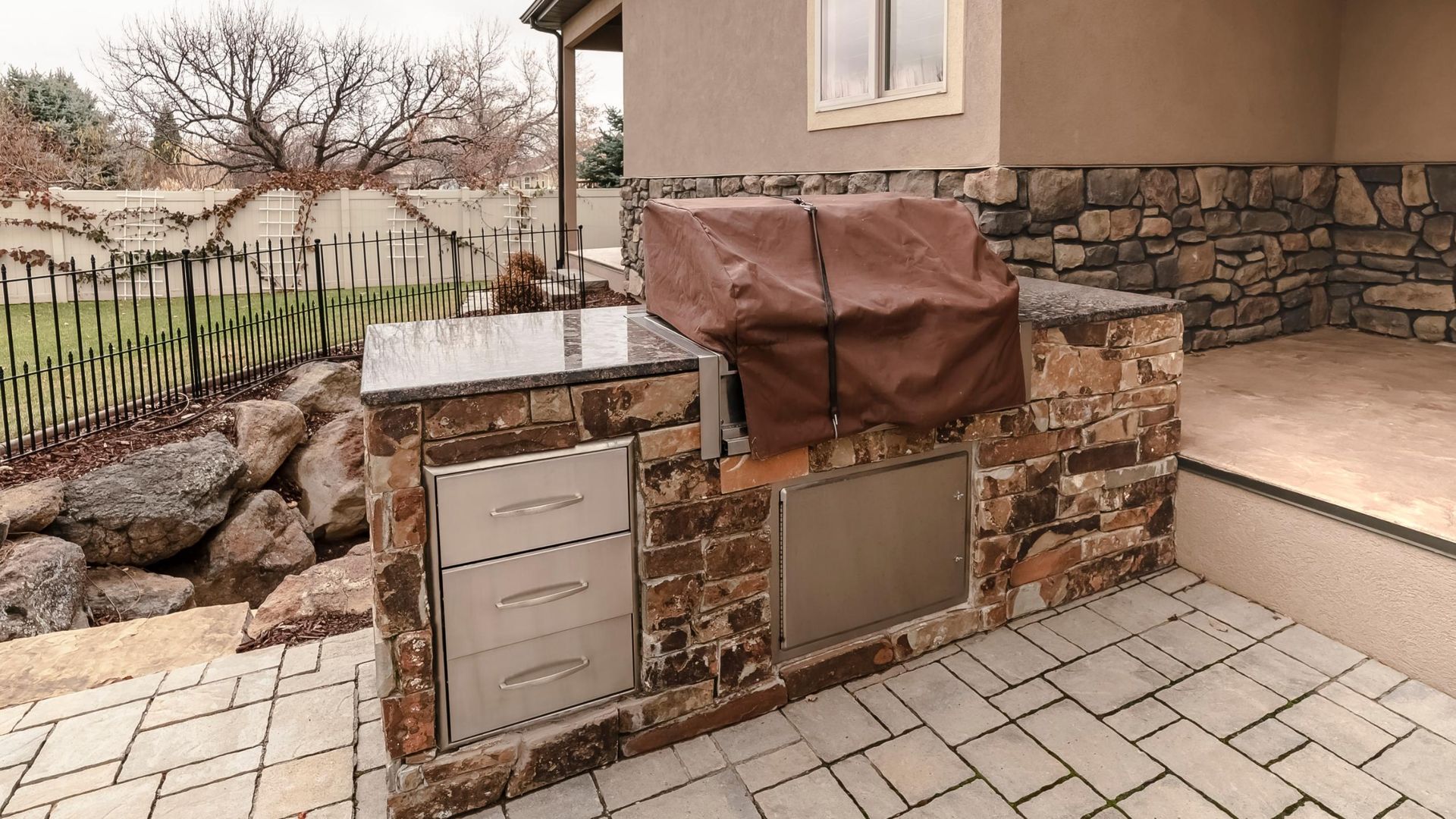Outdoor kitchen with stone facade, stainless steel appliances, and grill under cover.