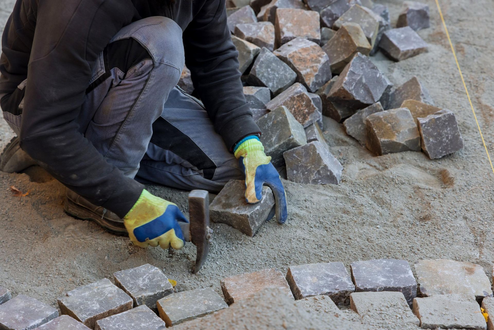 Construction worker laying cobblestones, using a hammer and wearing gloves, outdoors.