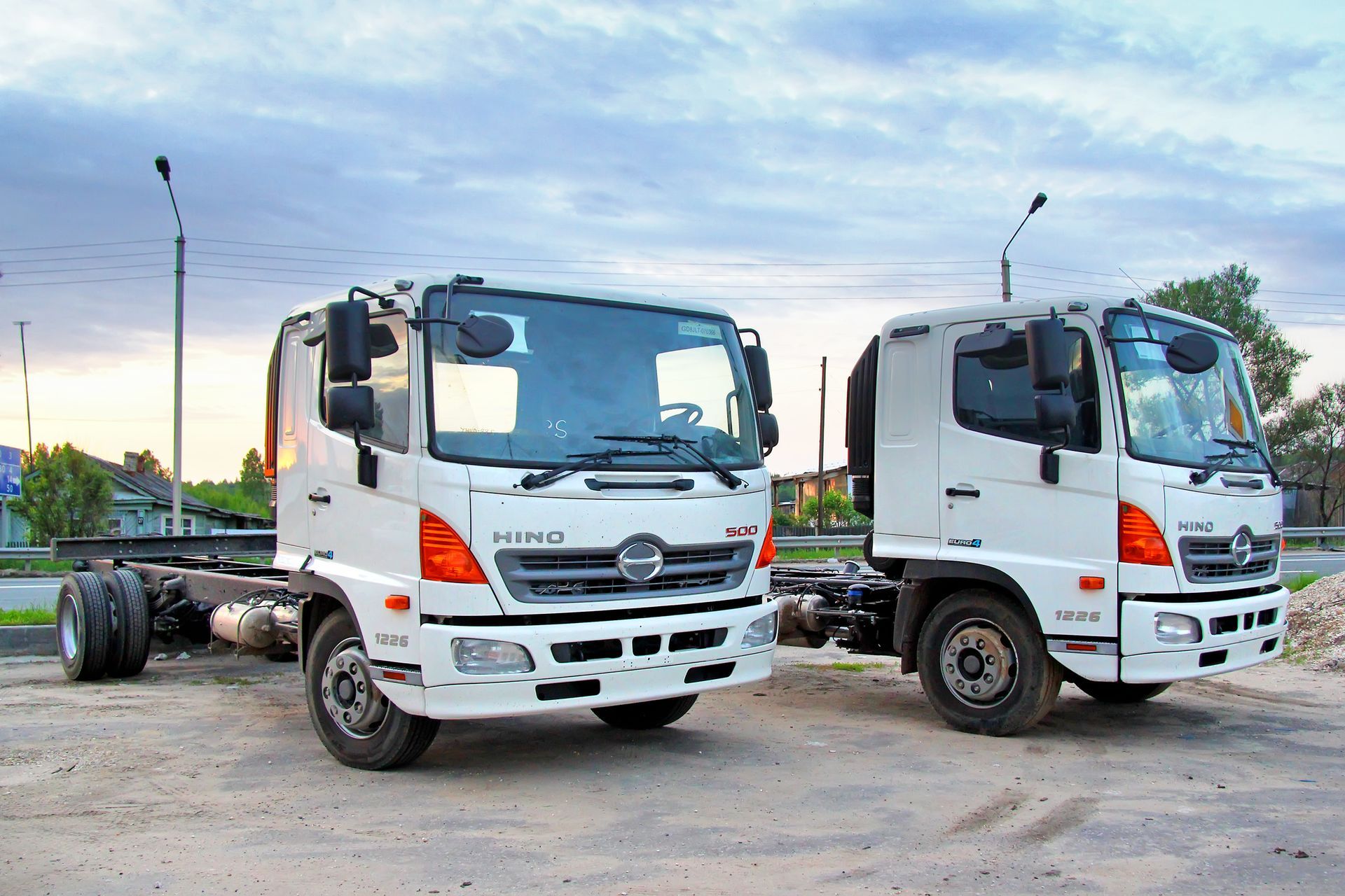 Two white Hino commercial truck chassis parked on a dirt lot against a cloudy sky at dusk.