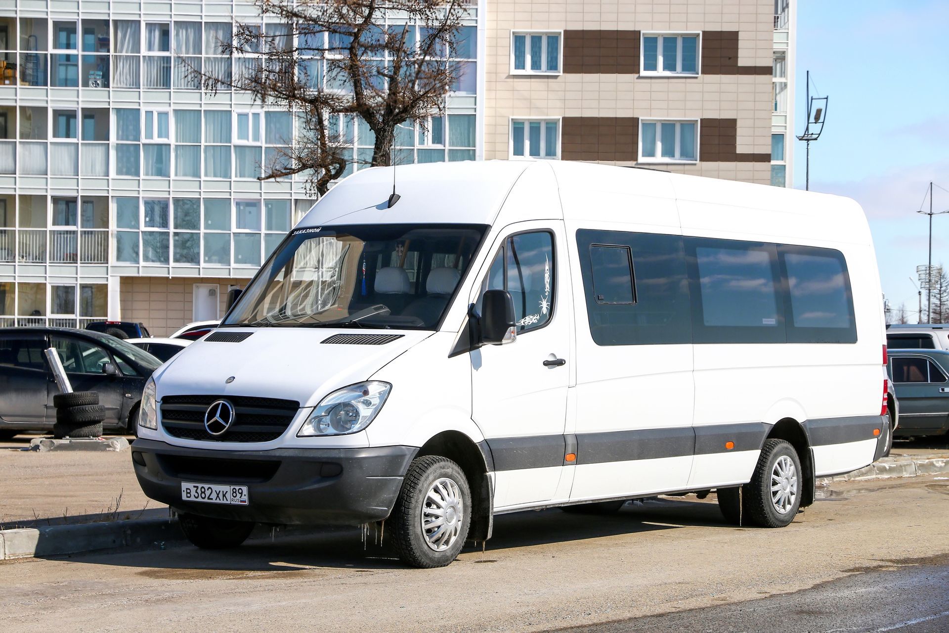 A white Mercedes-Benz Sprinter van parked on a gravel lot in front of a multi-story apartment building.