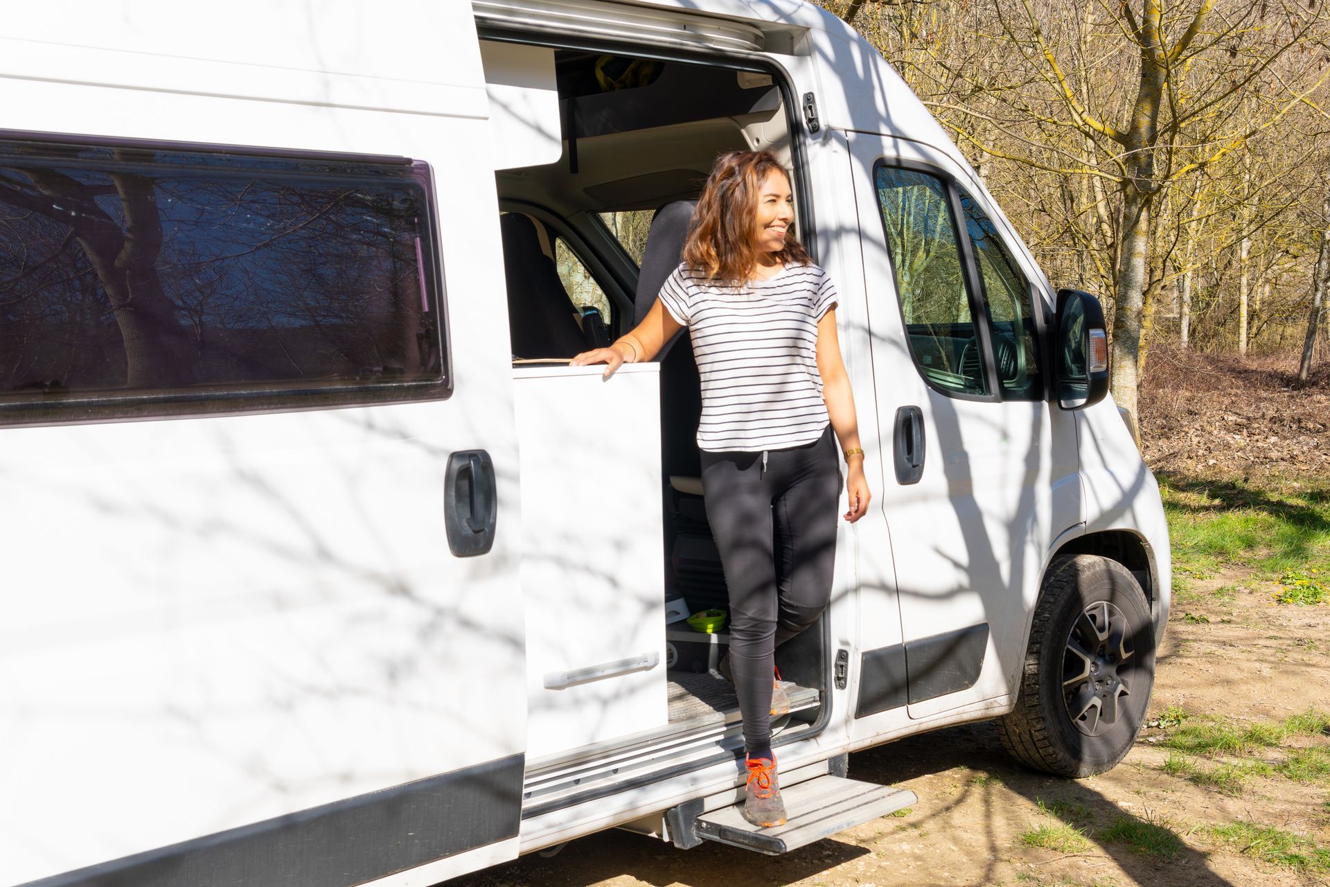 A smiling person stands in the open doorway of a white camper van, stepping onto a retractable footstool in a park.