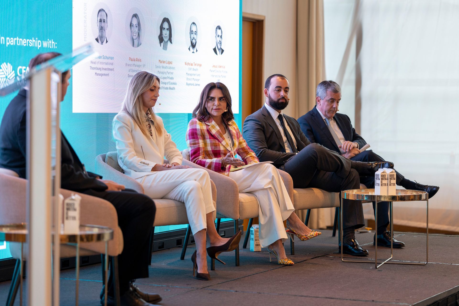 Panel discussion with five seated speakers on a stage, including a woman in white and a man in a suit.
