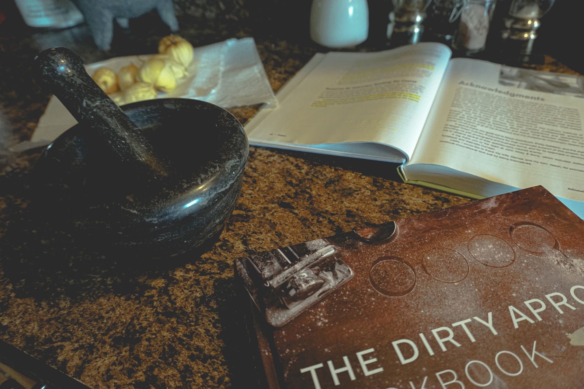 Mortar and pestle, cookbook, and ingredients on a countertop, possibly for a cooking project.