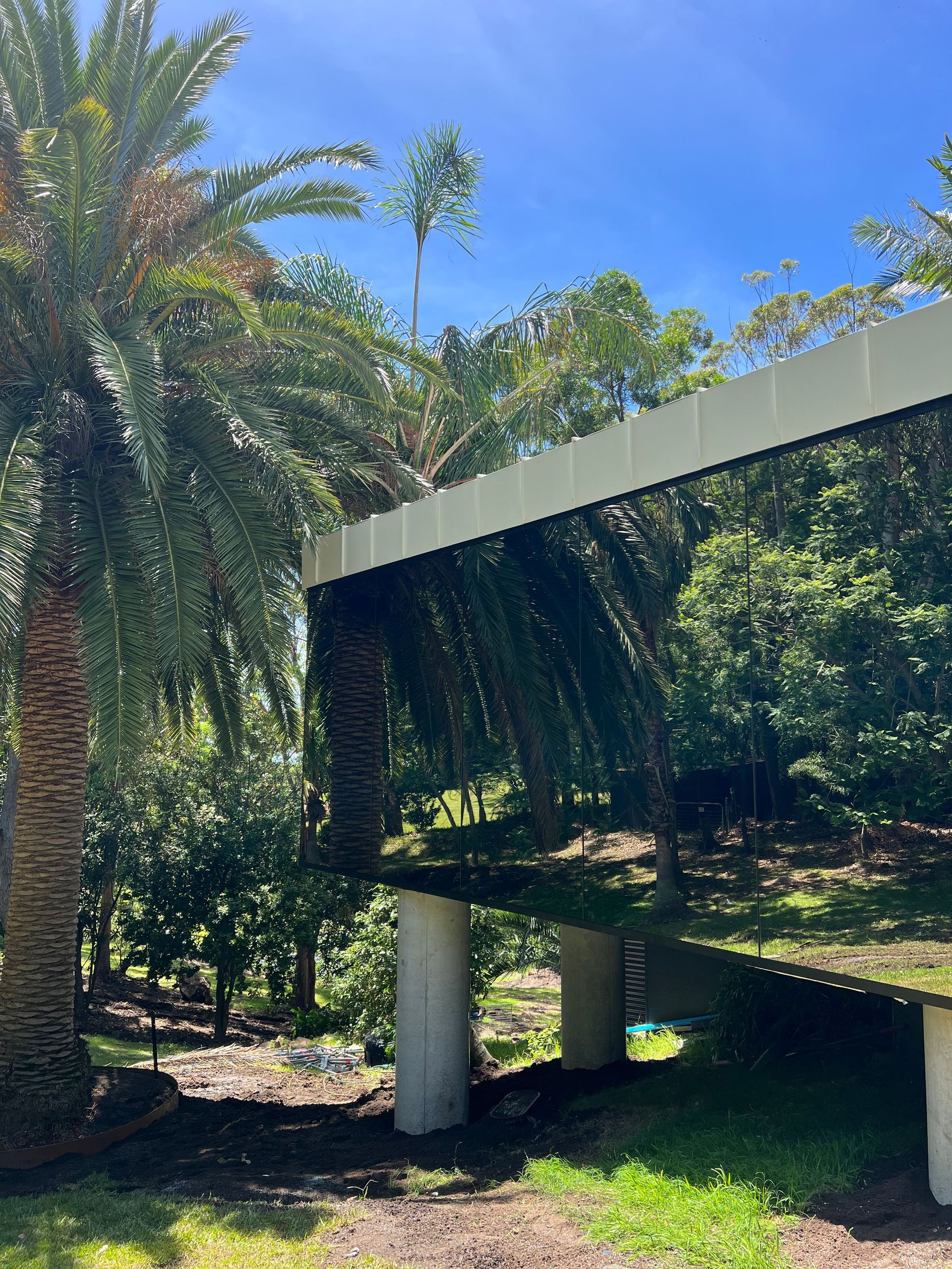 Mirror-like building reflects trees and sky; supported by white columns, palm tree on left, grassy area.