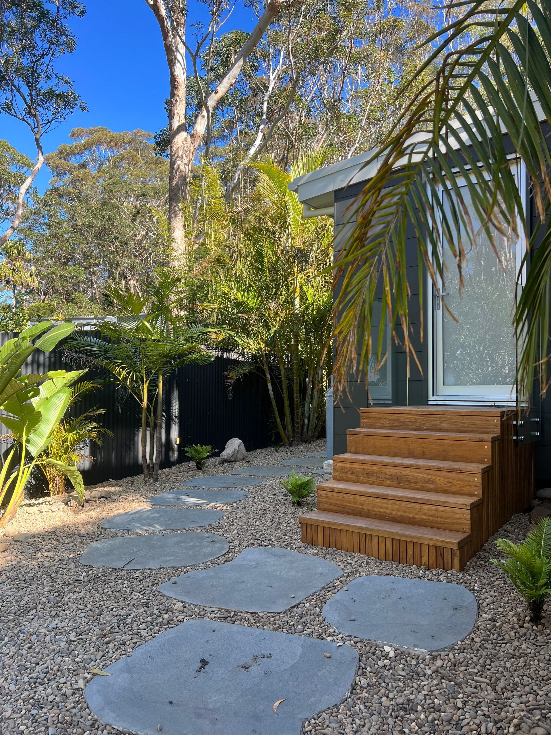 Stone path leads to wooden steps and a doorway of a green cabin, surrounded by lush greenery.