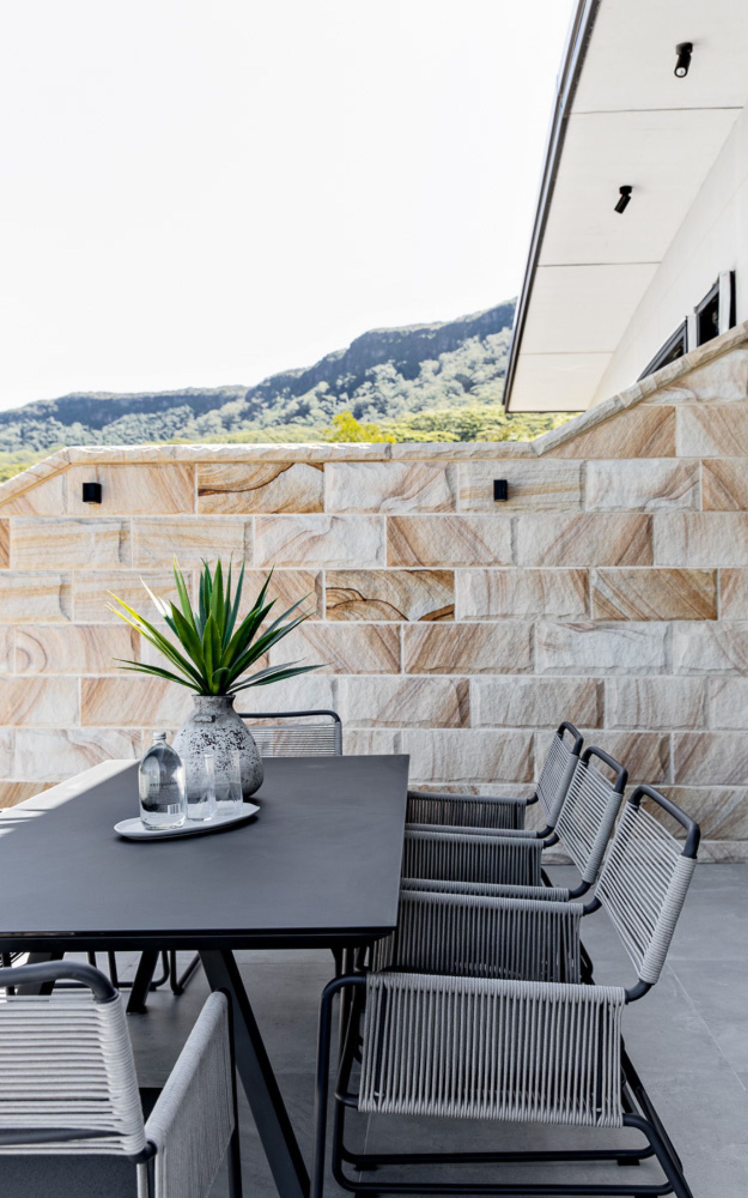 Outdoor dining area with grey table and chairs, against a stone wall and mountain view — LG Constructions in Thirroul, NSW