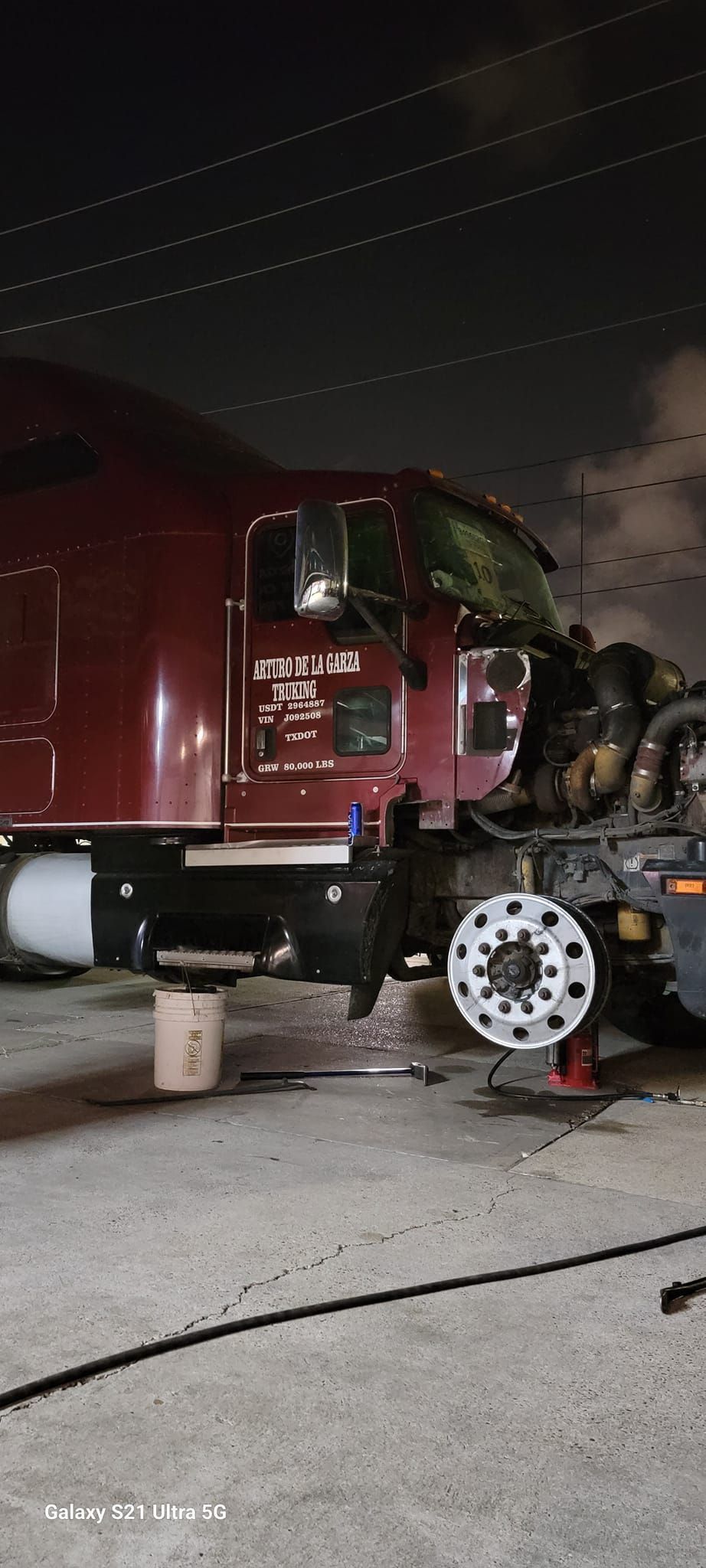A maroon semi-truck has a tire removed at night. An open jack holds the vehicle up near the wheel.