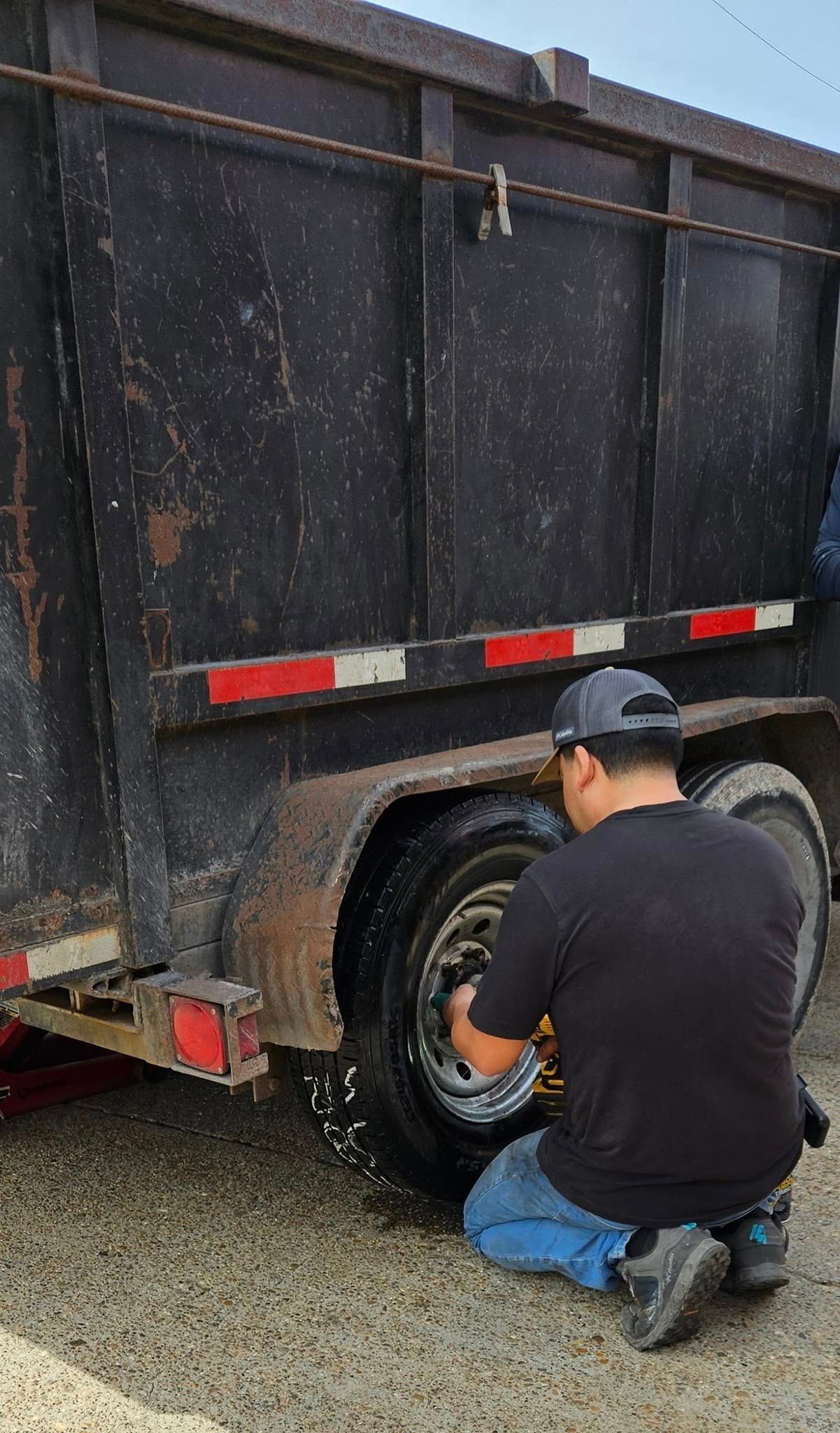 Person kneeling by a trailer tire, appears to be inflating it outdoors.