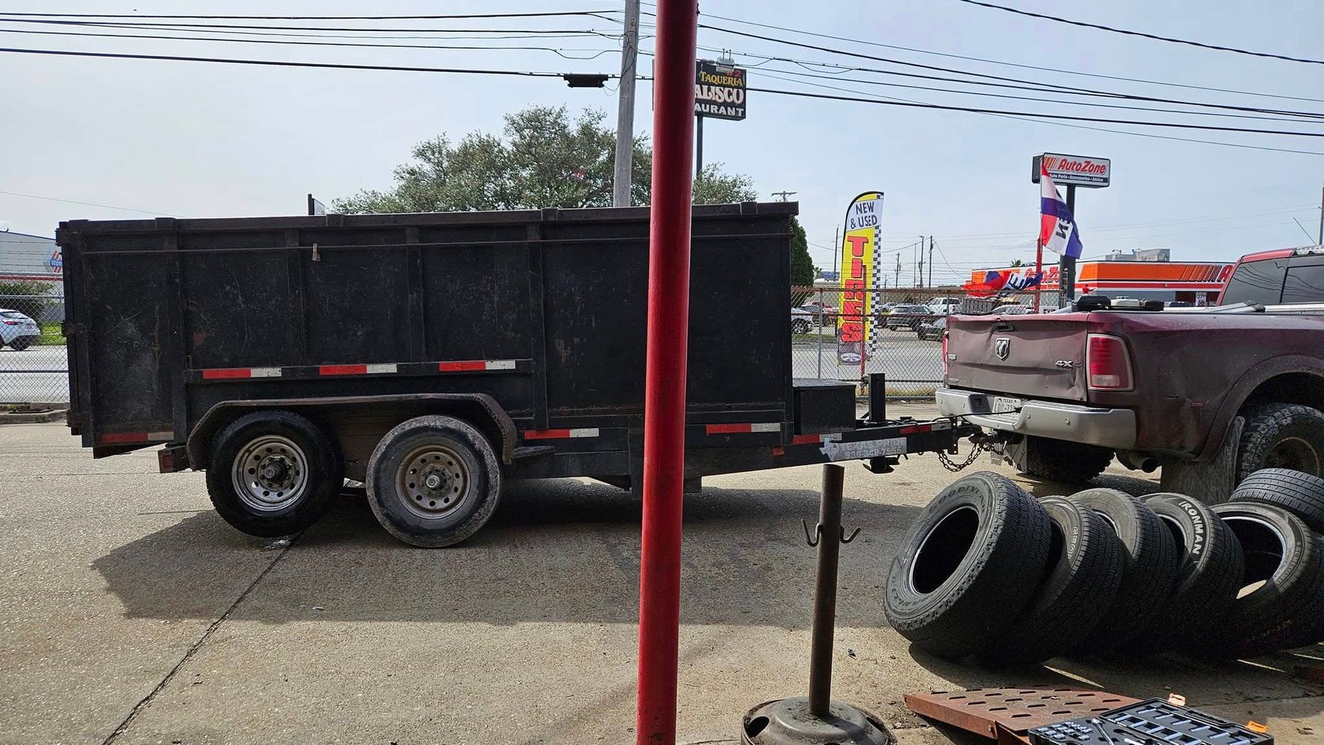 Black dump trailer hitched to a maroon pickup truck, parked in front of a shop with tires and a sign.