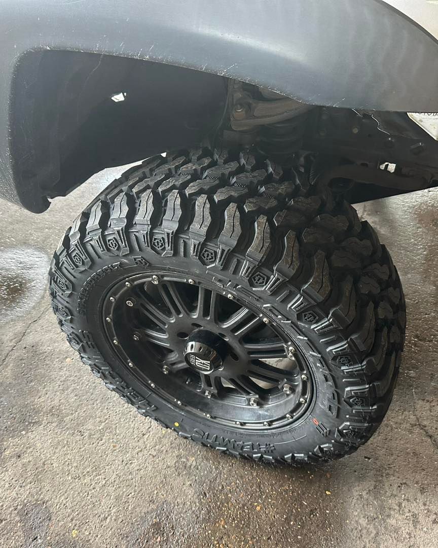 Black off-road tire on a black rim, under the fender of a truck on a concrete surface.