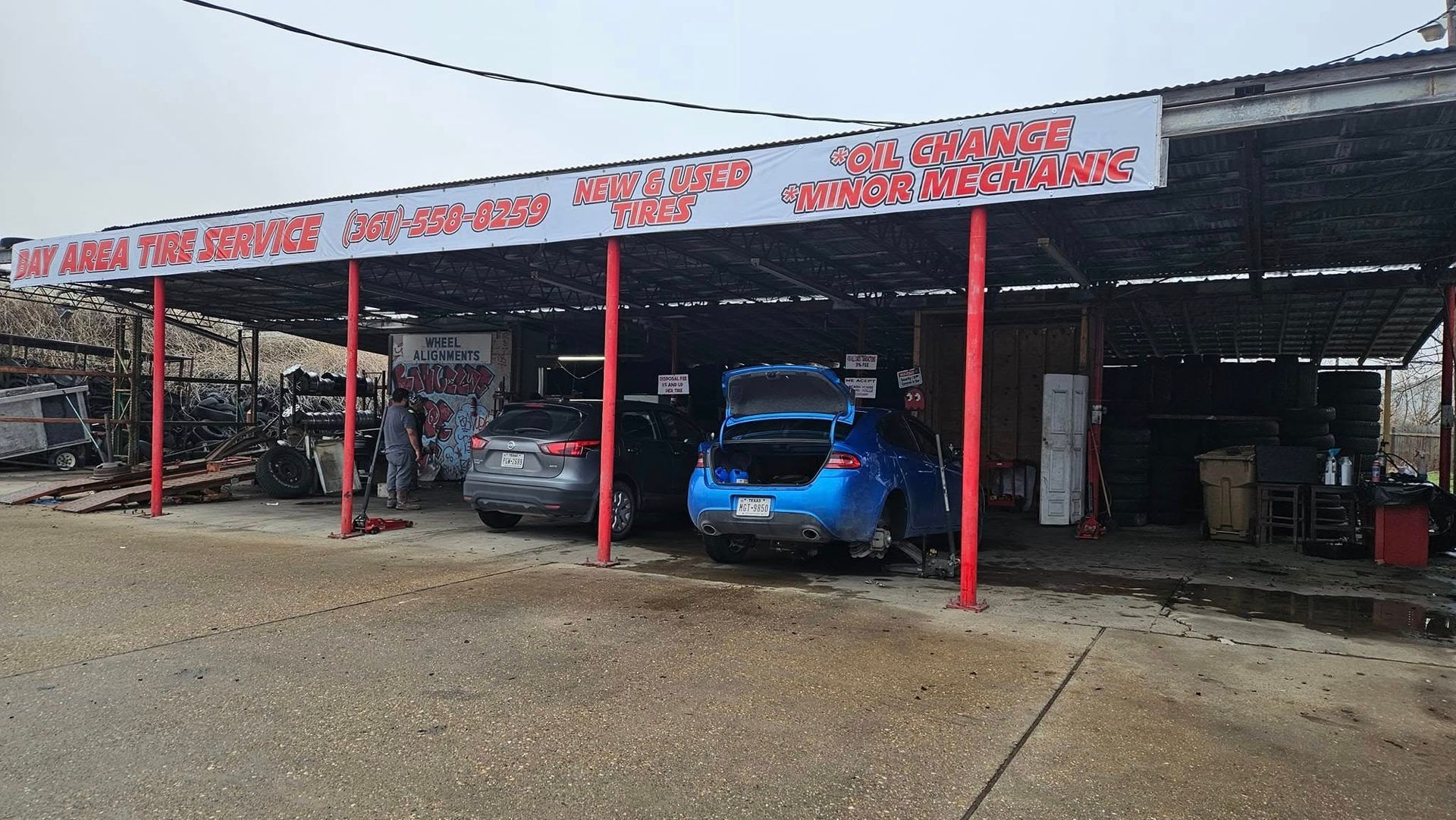 A car repair shop with several vehicles under a covered area, red pillars, and a sign.