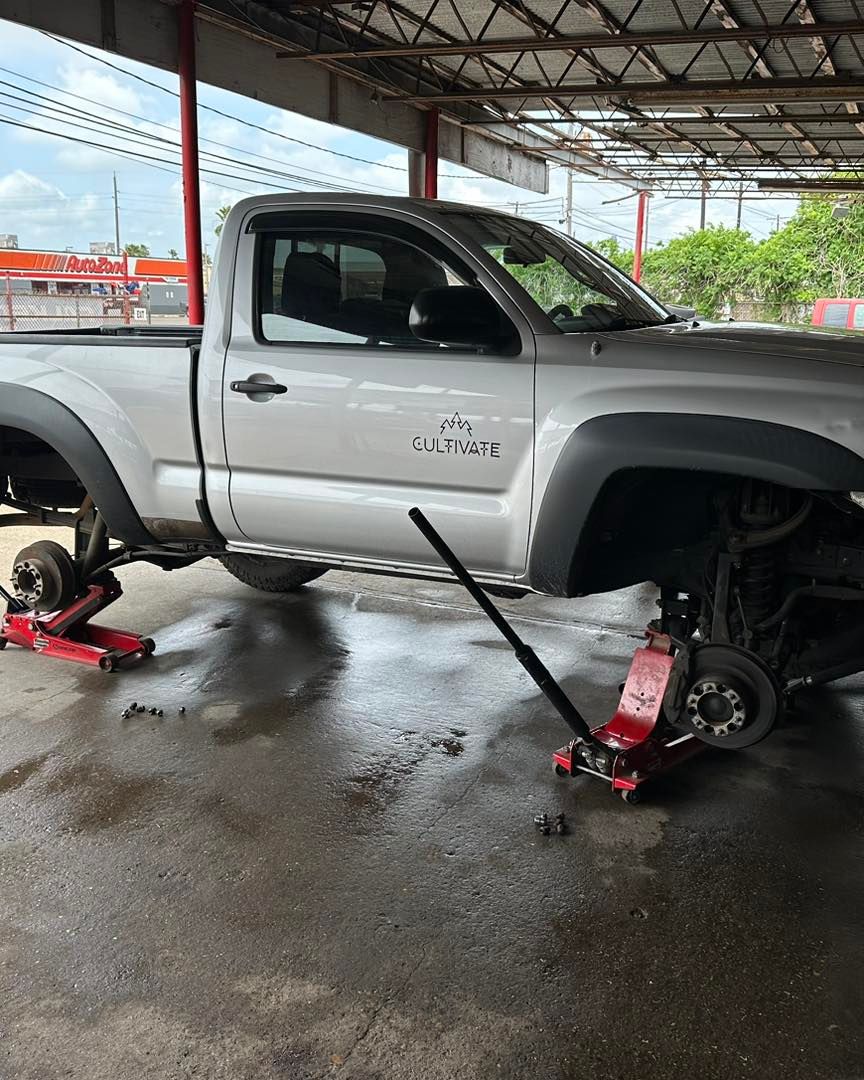 Silver Toyota truck raised on jacks, wheels removed, under a metal roof at a repair shop.