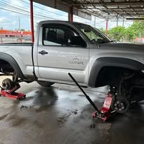 Silver pickup truck on jack stands in a repair shop, with tools and a wet floor.