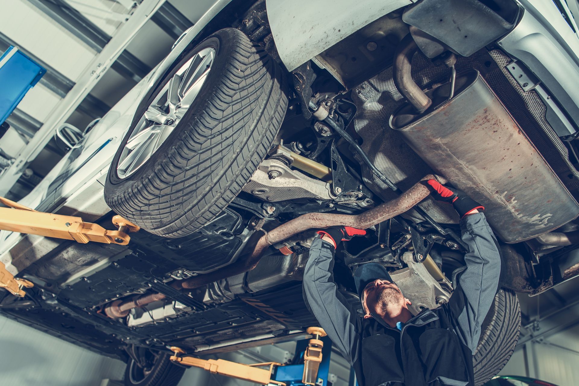 Mechanic working on the underside of a car in a garage, using tools near exhaust system.