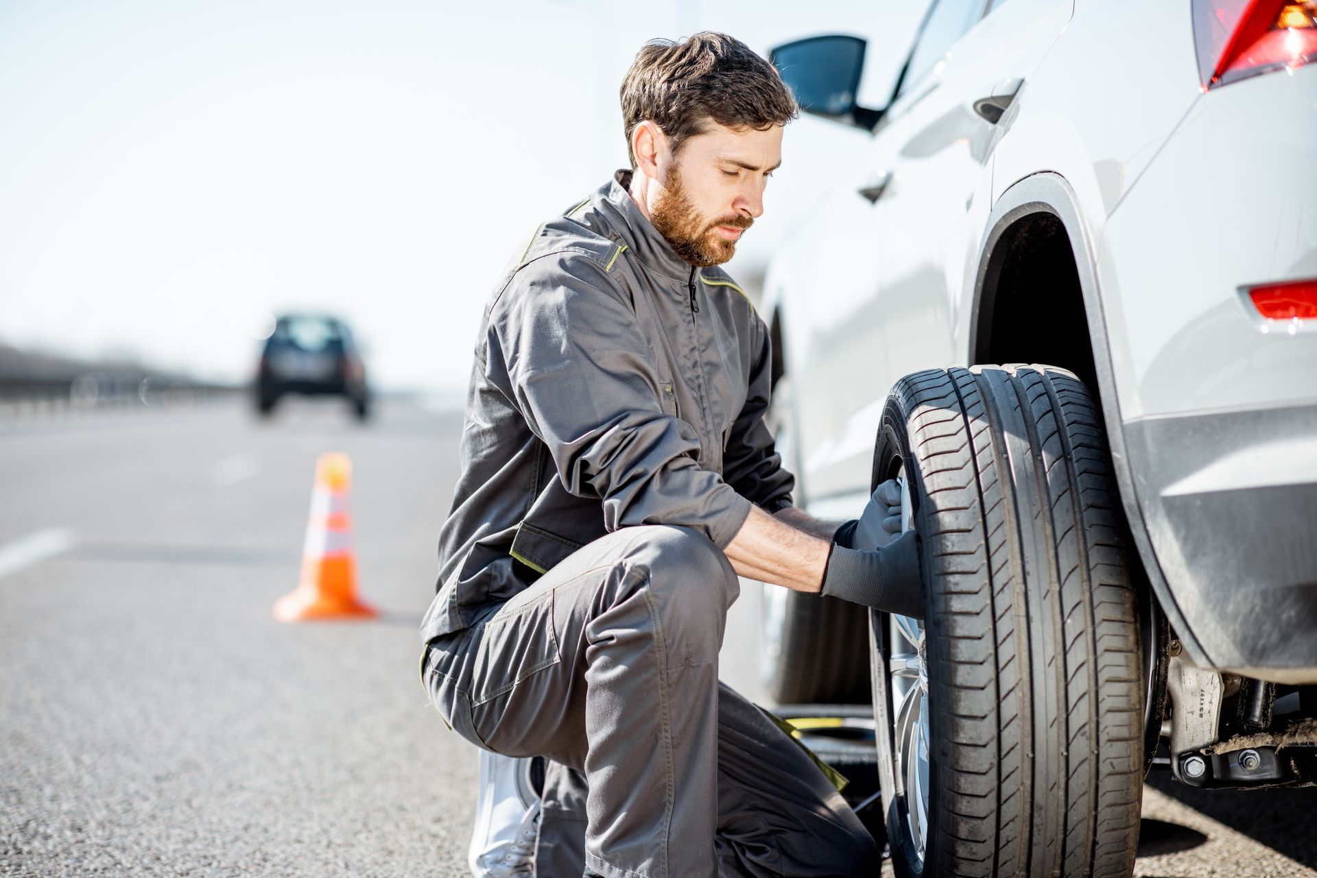 Man changing a tire on a white car on the side of a highway. Orange cone in the background. Man changing a tire on a white car on the side of a highway. Orange cone in the background.