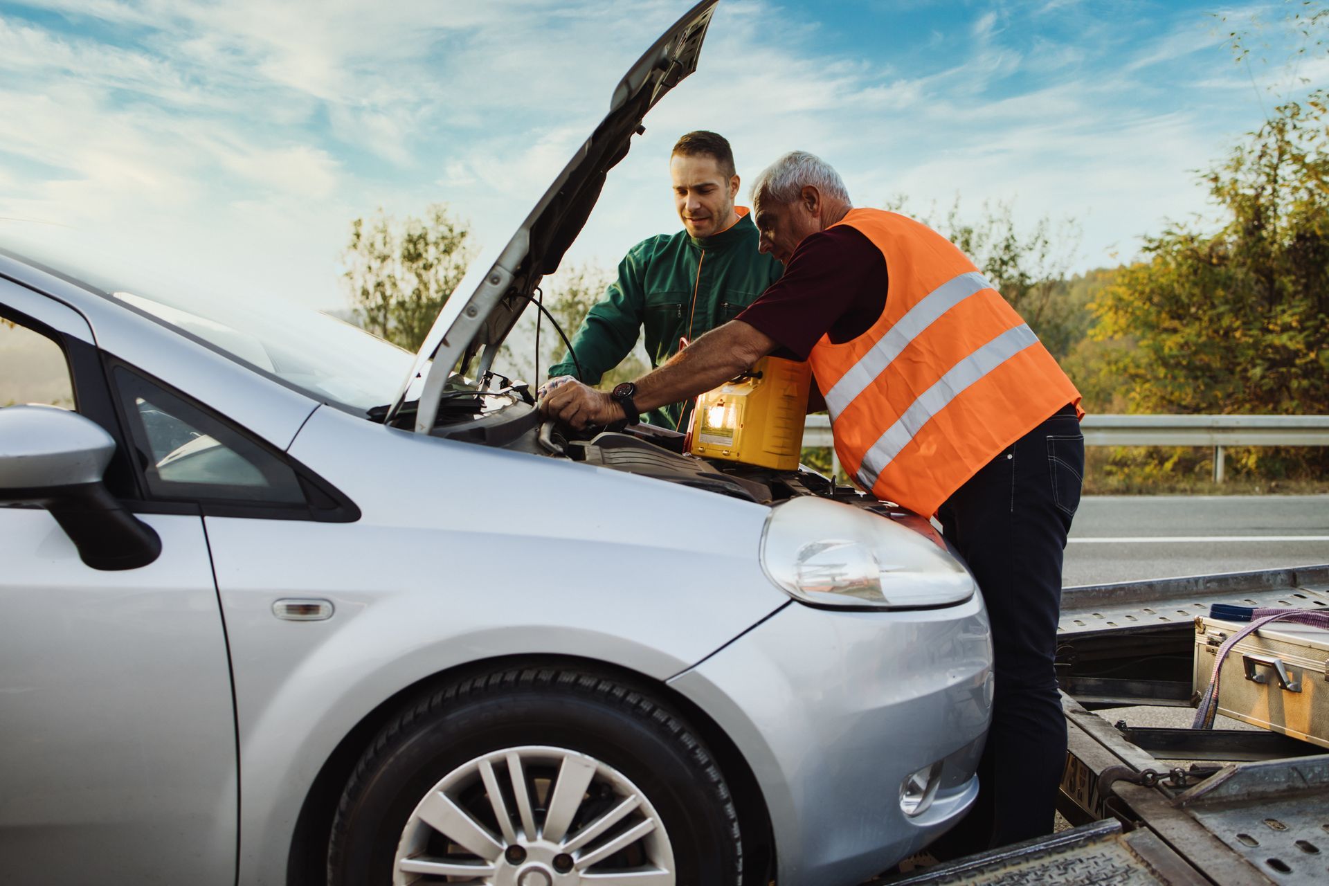 Two men by a silver car with the hood up, roadside; one in an orange vest pours liquid. Two men by a silver car with the hood up, roadside; one in an orange vest pours liquid.