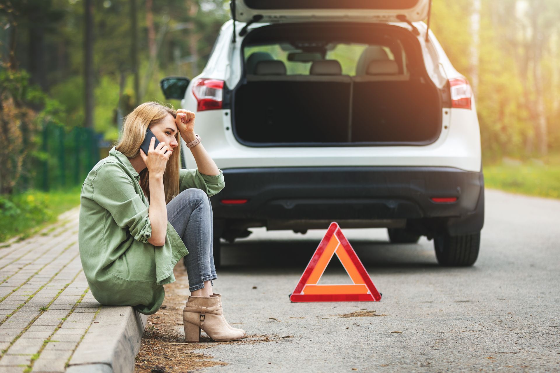 Woman on phone by broken-down car, sitting near a road with a warning triangle. Woman on phone by broken-down car, sitting near a road with a warning triangle.