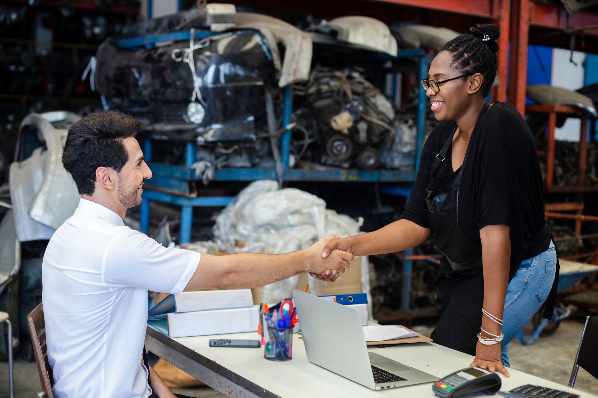 Man and woman shaking hands over a desk in a cluttered garage, smiling. Laptop and boxes present. Man and woman shaking hands over a desk in a cluttered garage, smiling. Laptop and boxes present.