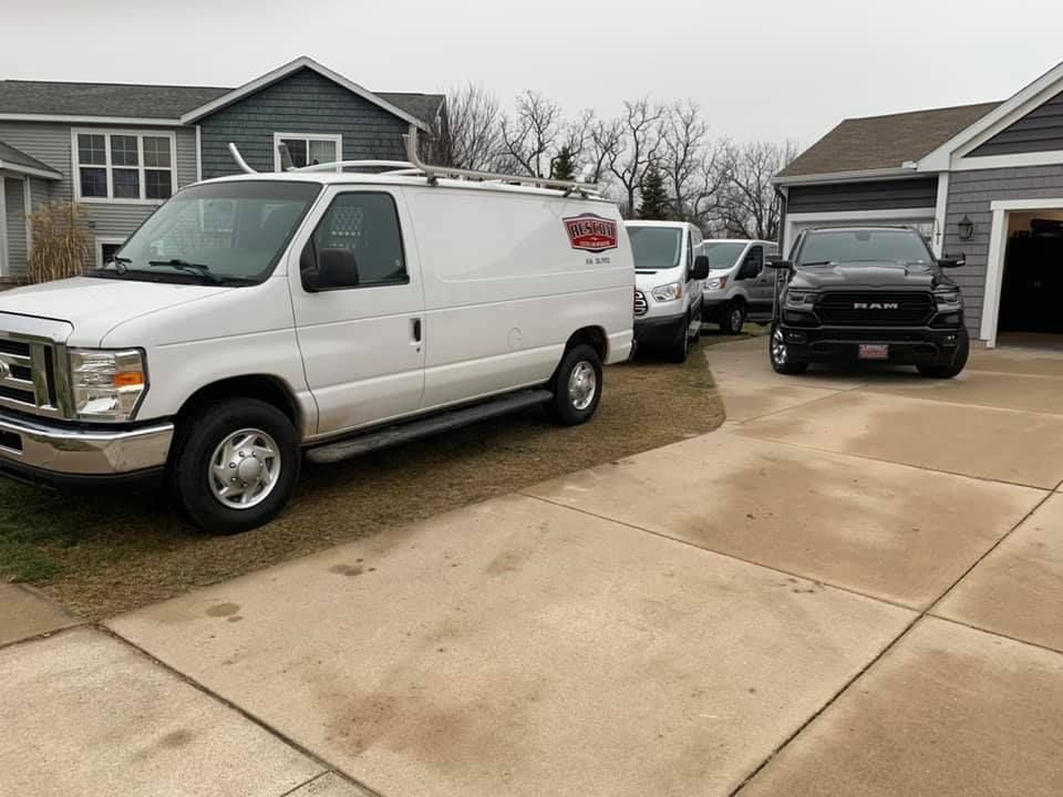 A row of white vans are parked in a driveway in front of a house.
