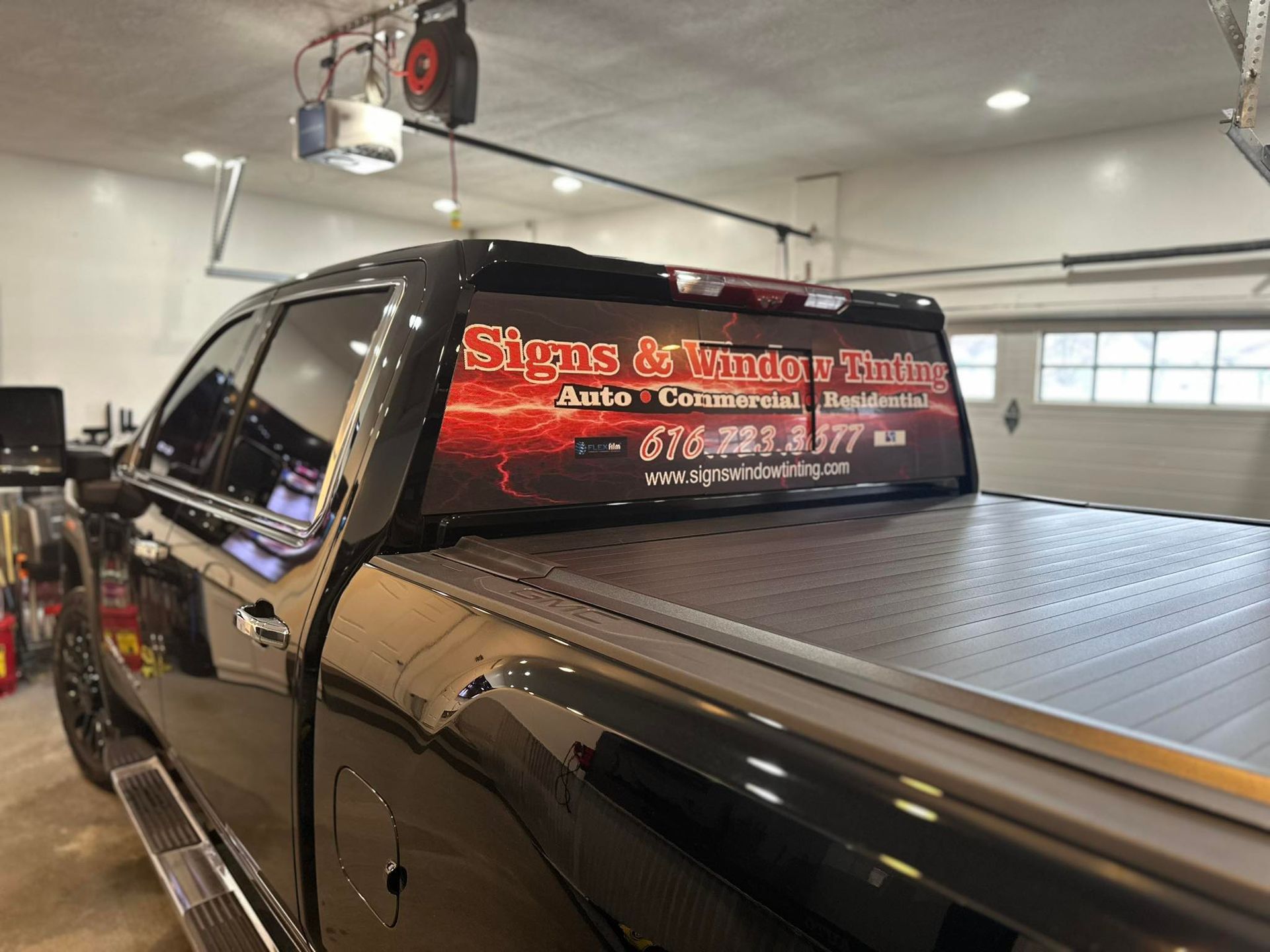 A black truck with a red sign on the back window is parked in a garage.