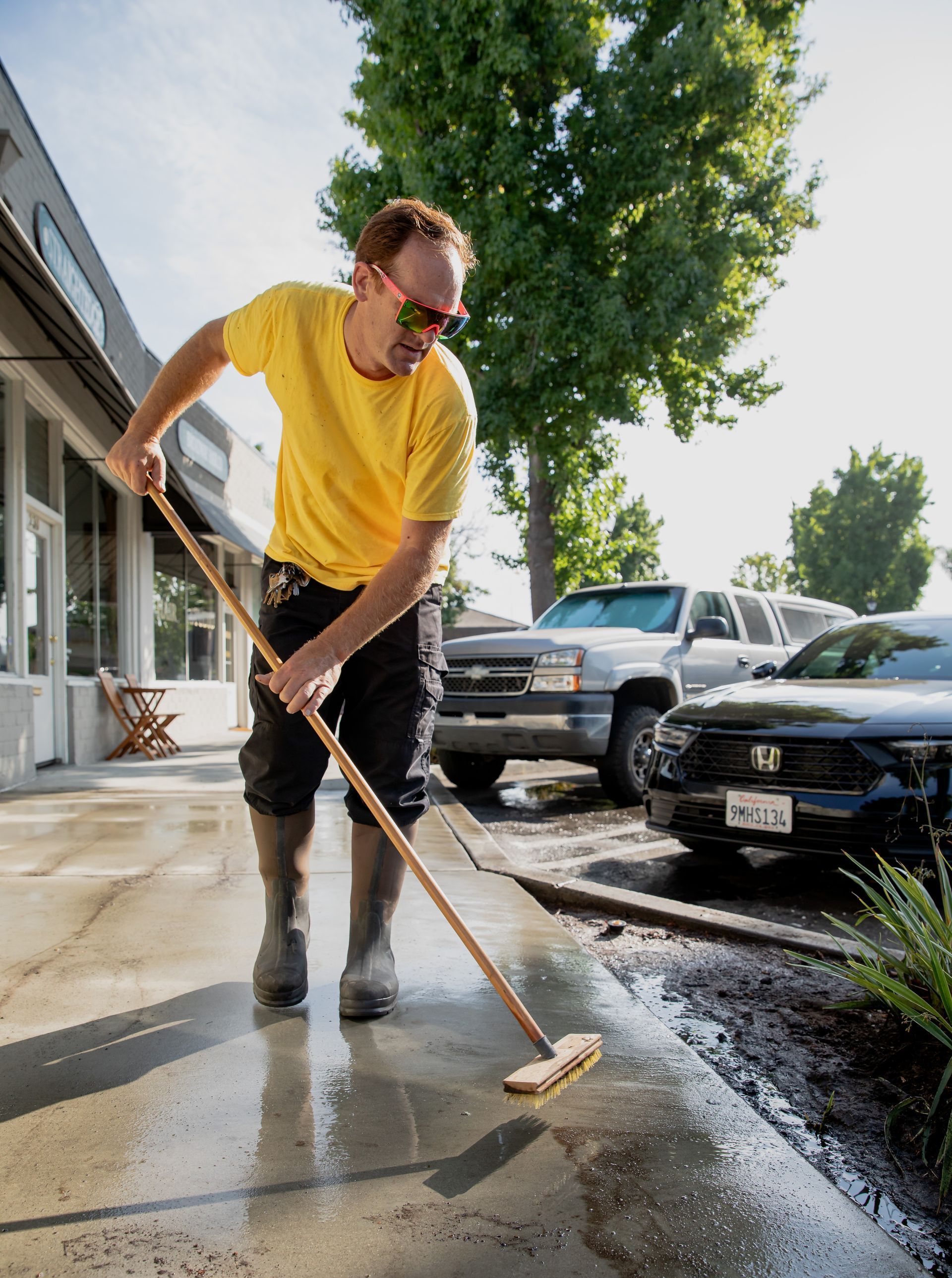 A man in a yellow shirt is cleaning the sidewalk with a broom.