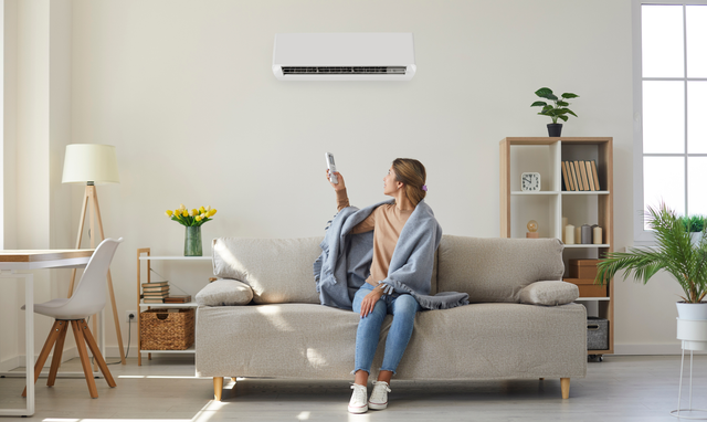 A person wrapped in a blanket sits on a sofa, aiming a remote control at a wall-mounted air conditioner in a bright room.