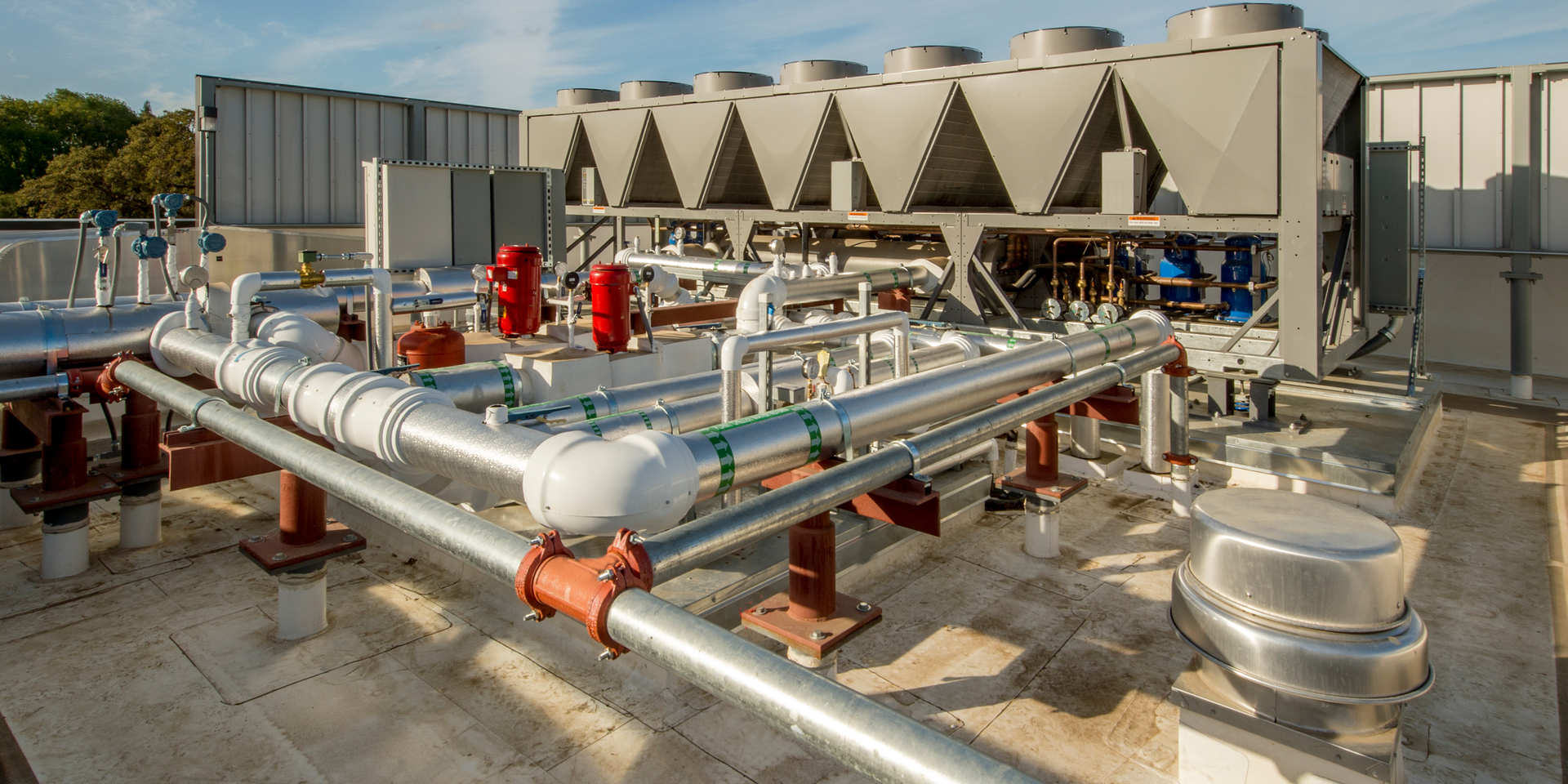 Industrial HVAC chiller unit with silver piping and red valves on a flat rooftop against a blue sky.