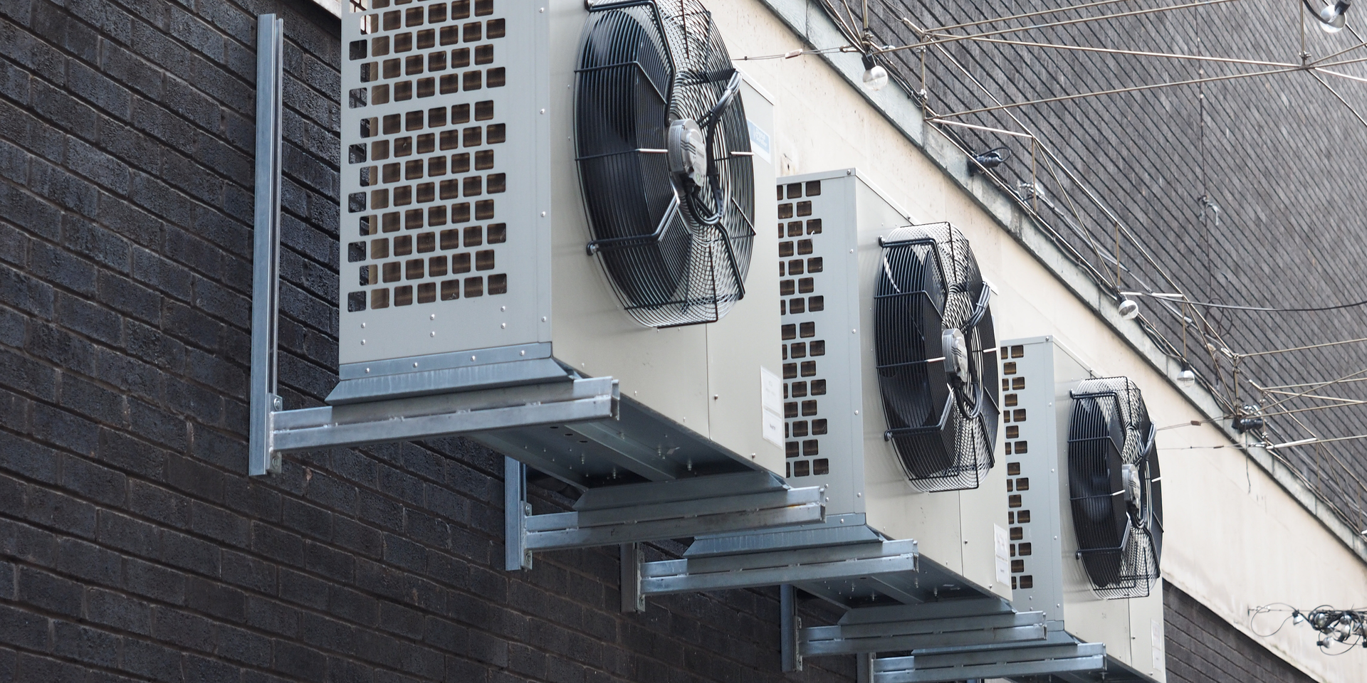 Three beige industrial air conditioning units mounted in a row on a dark brick wall.