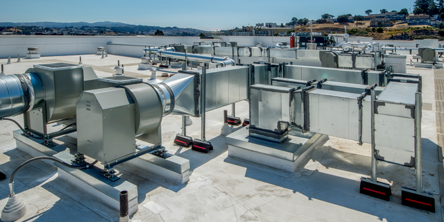 HVAC equipment and silver metal ductwork installed on a flat commercial building rooftop under a bright blue sky.