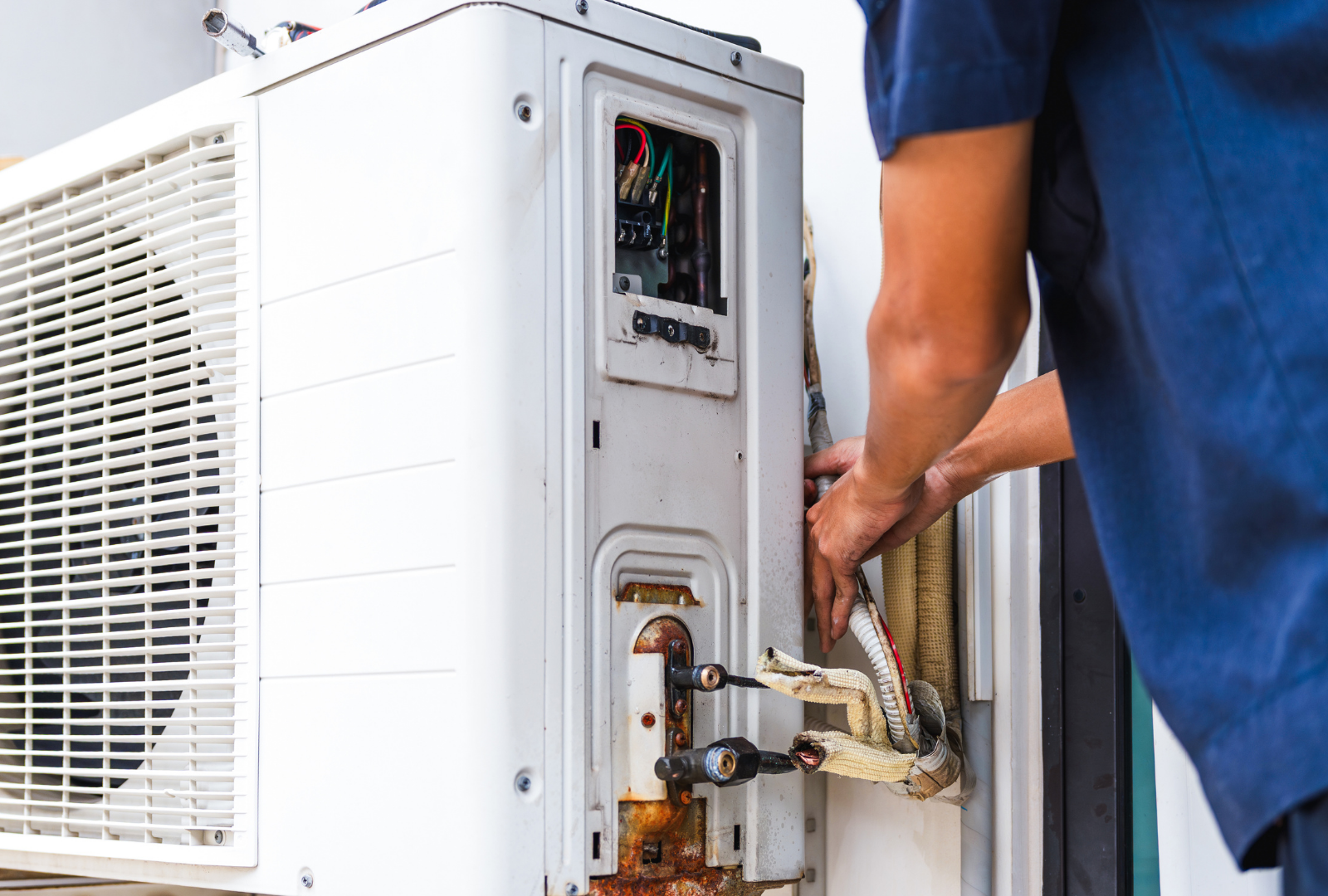 A technician in a blue uniform repairs the open wiring and pipe connections of an outdoor air conditioning unit.