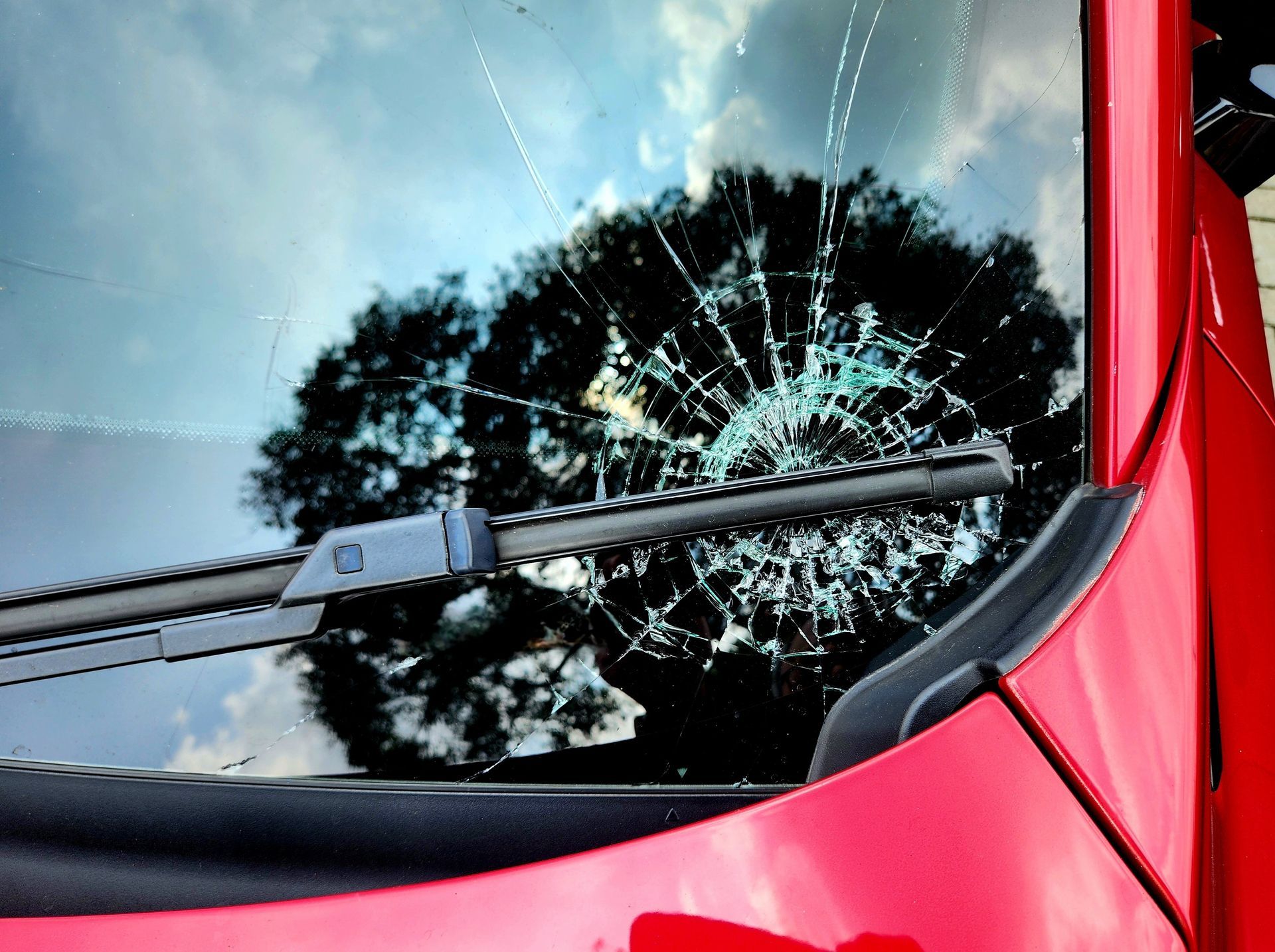 Cracked windshield on a red car with a tree reflection and visible wiper blade