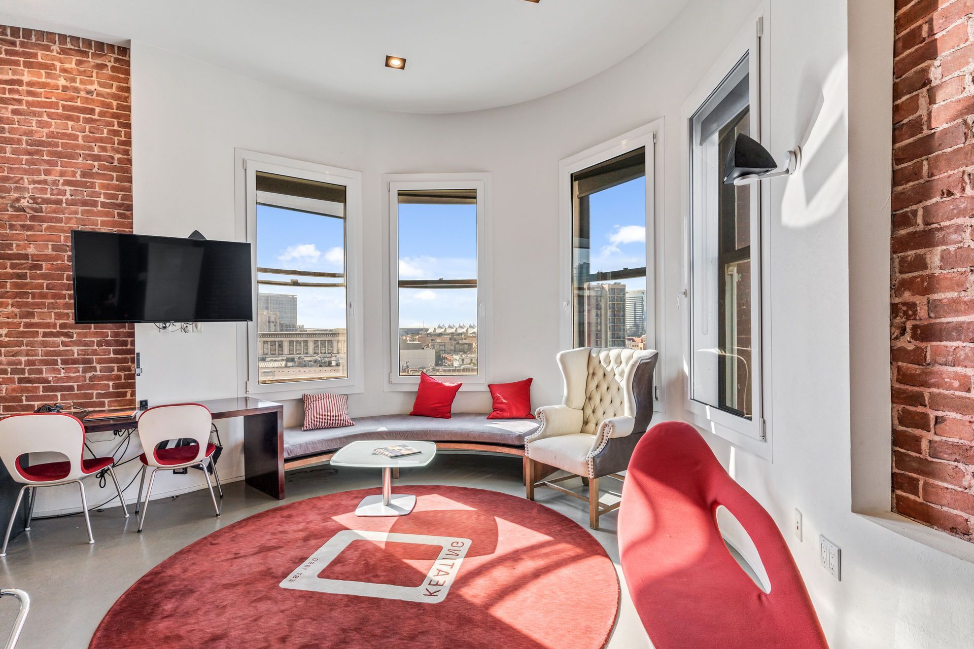 Bright living room with brick walls, curved windows, red rug, and chairs.