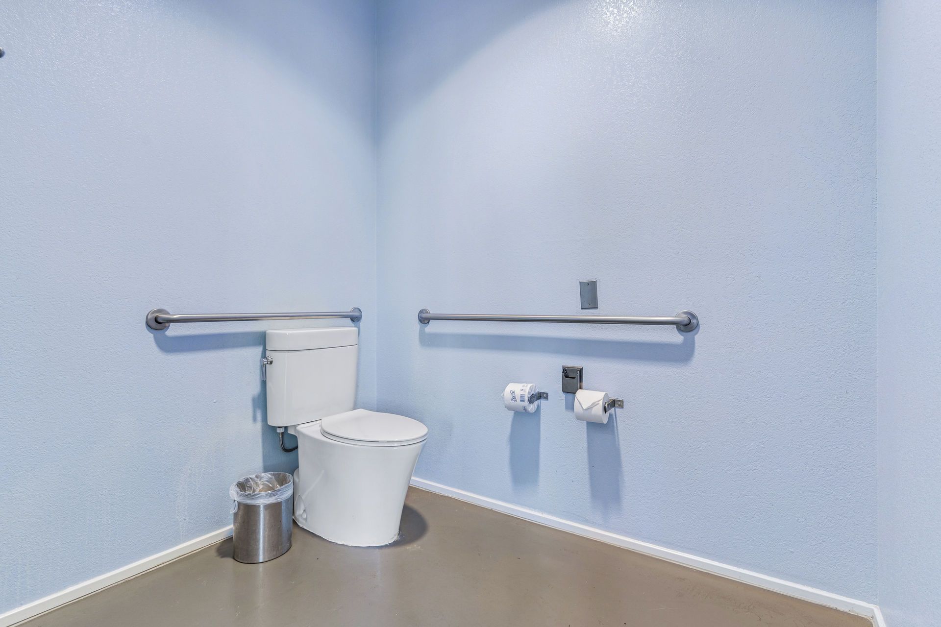Accessible bathroom with grab bars, toilet, and paper towel dispenser. Blue walls and neutral floor.