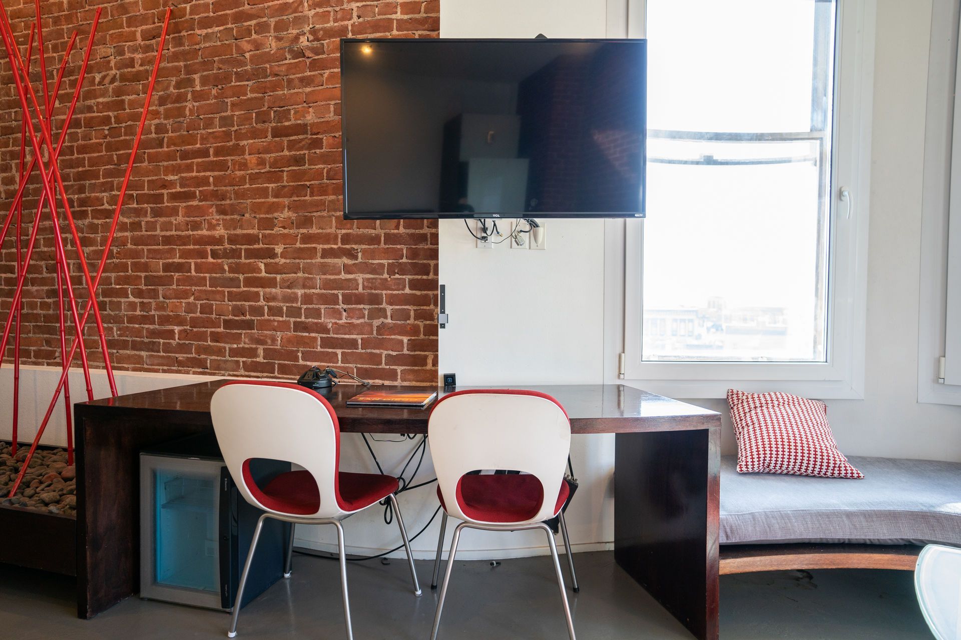 A room with a brick wall, a desk with two chairs, a TV, and a window.