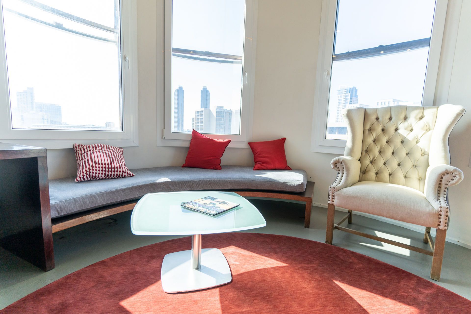 Cozy room with curved seating, red pillows, an armchair, and a circular rug. Bright windows offer a city view.