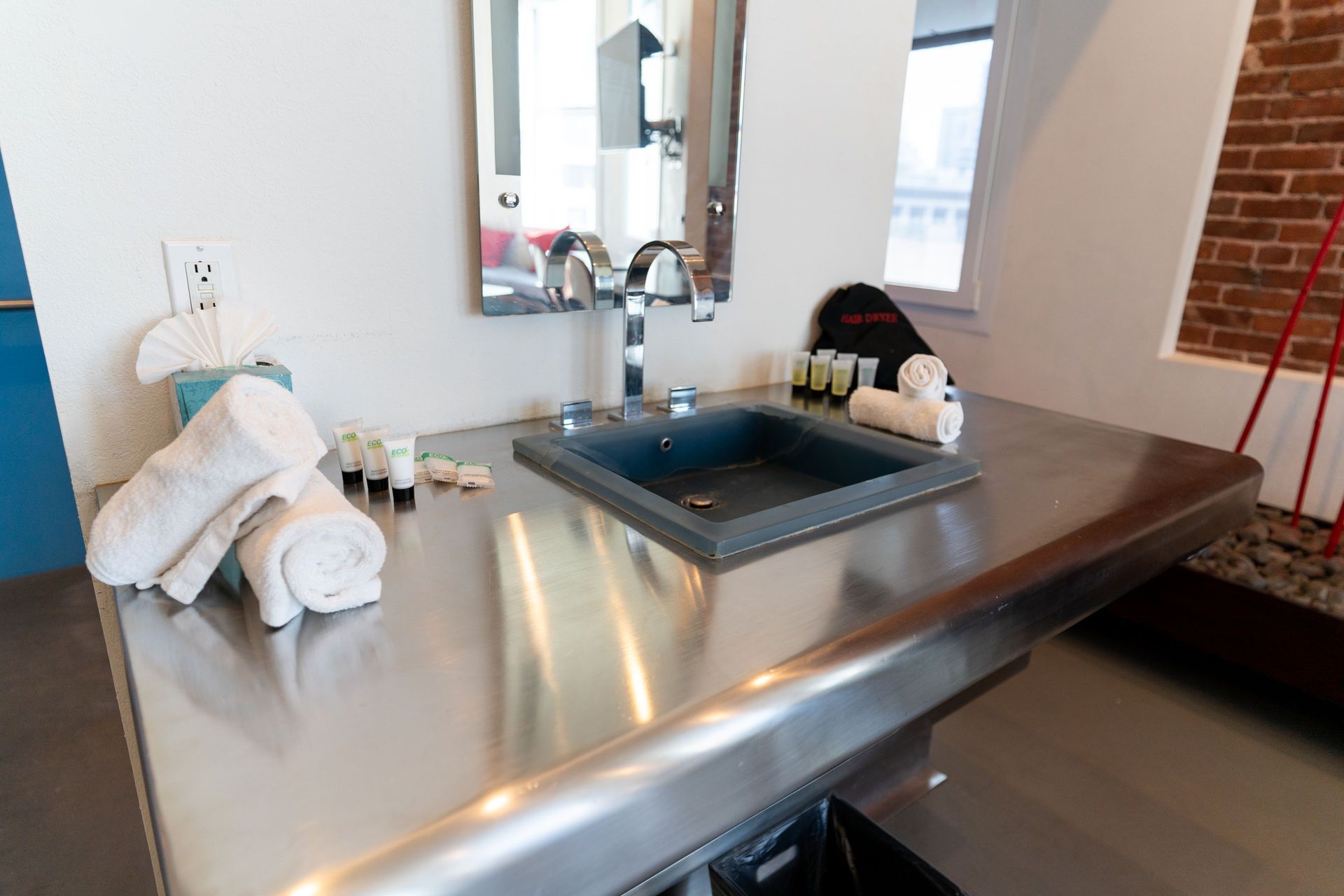 Bathroom with stainless steel counter, gray sink, mirror, towels, toiletries, and brick wall.