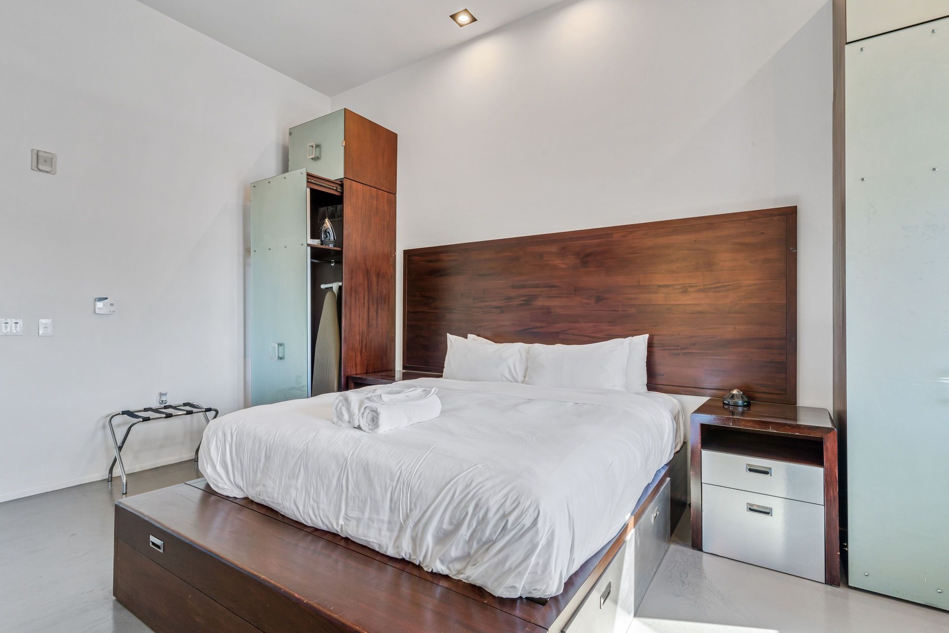 Bedroom with a bed, nightstand, closet, and a small table; white walls and a wood headboard.
