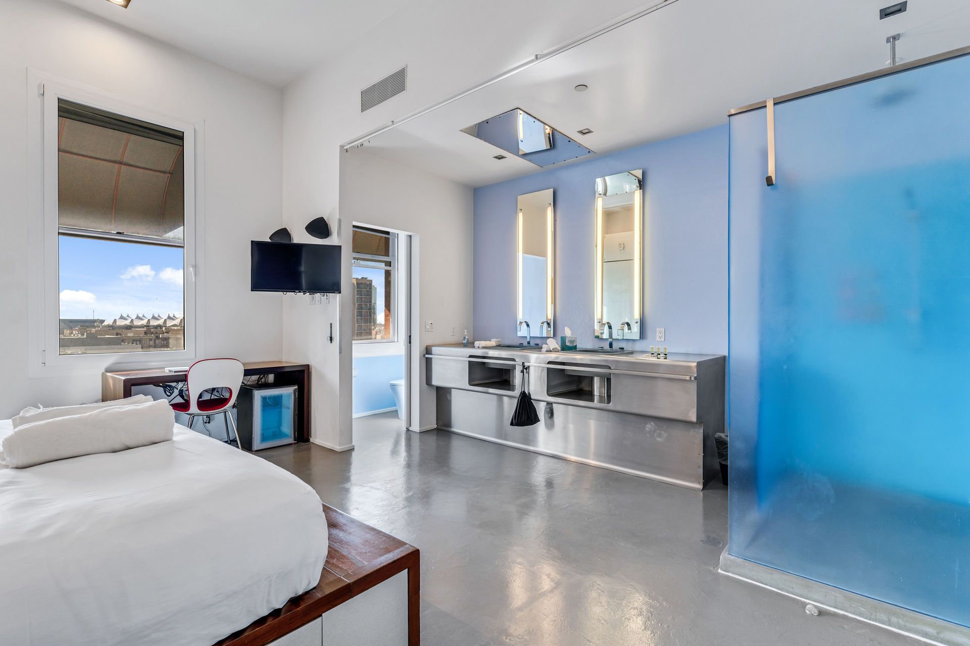 Modern bathroom with double sinks, blue shower, and bed. Bright, white walls, gray floor.