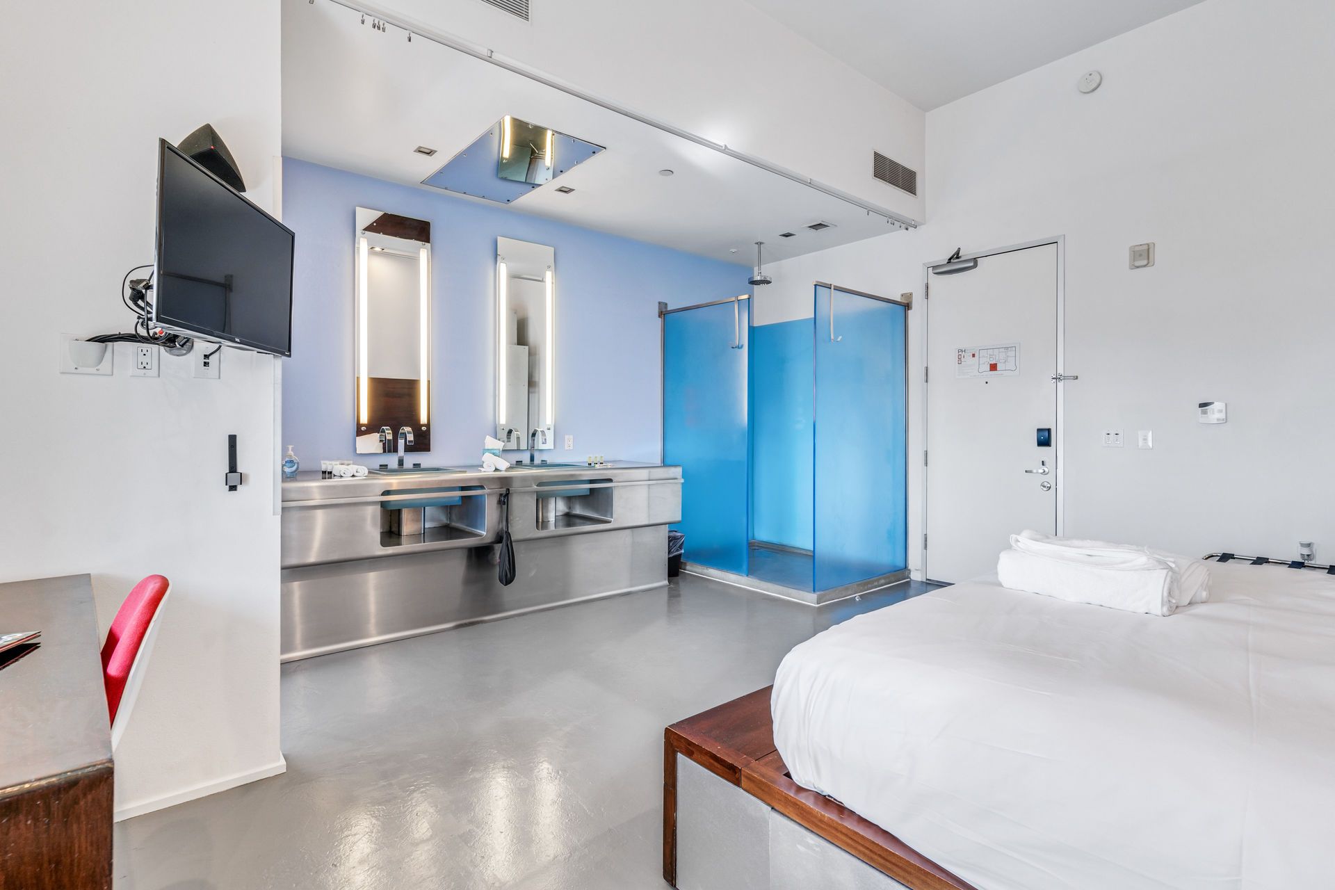 Modern hotel room with a bed, stainless steel sink, blue-walled shower, and TV.