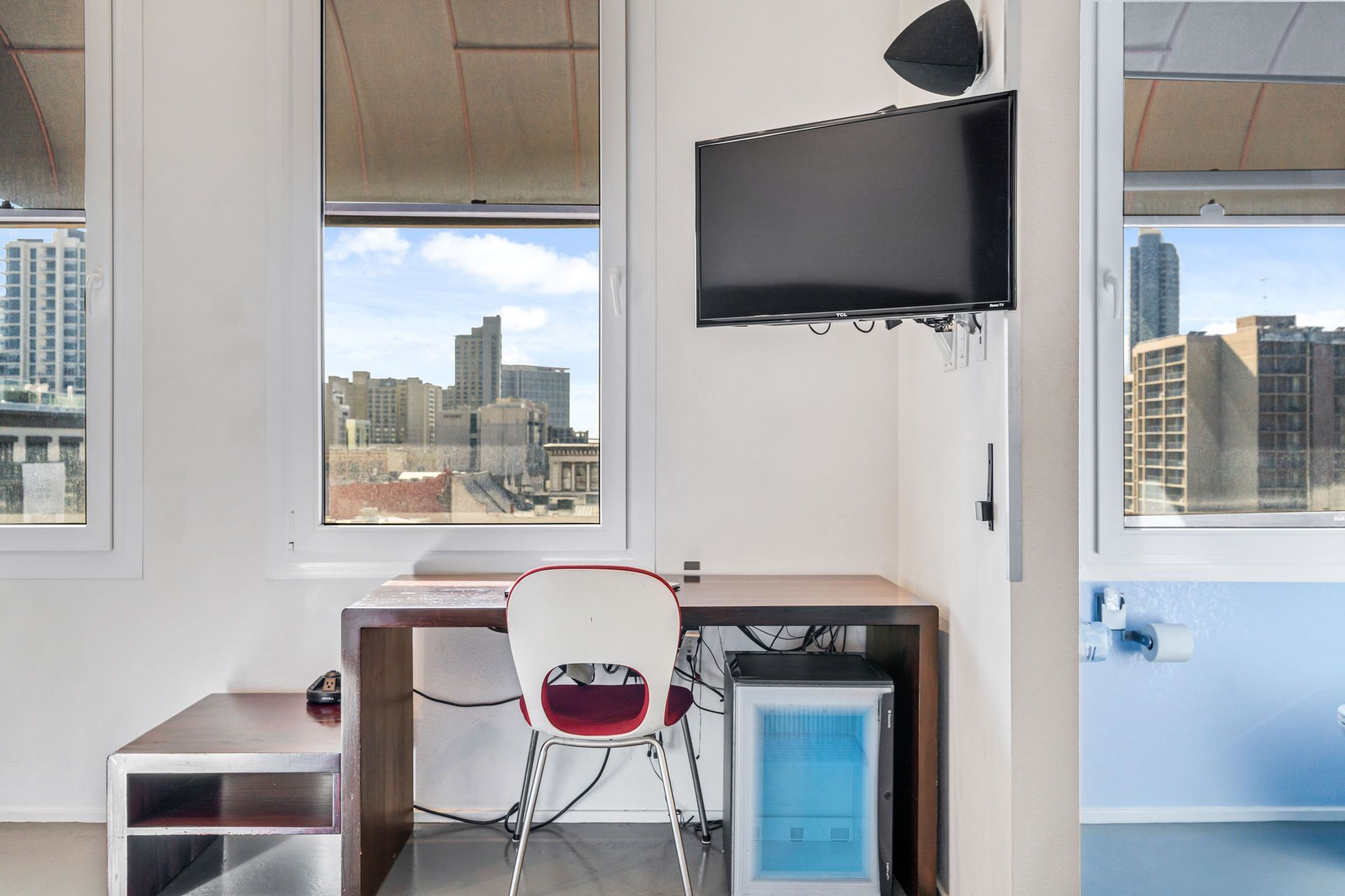 Desk area with window view of city buildings, chair, and small refrigerator. TV mounted on the wall.