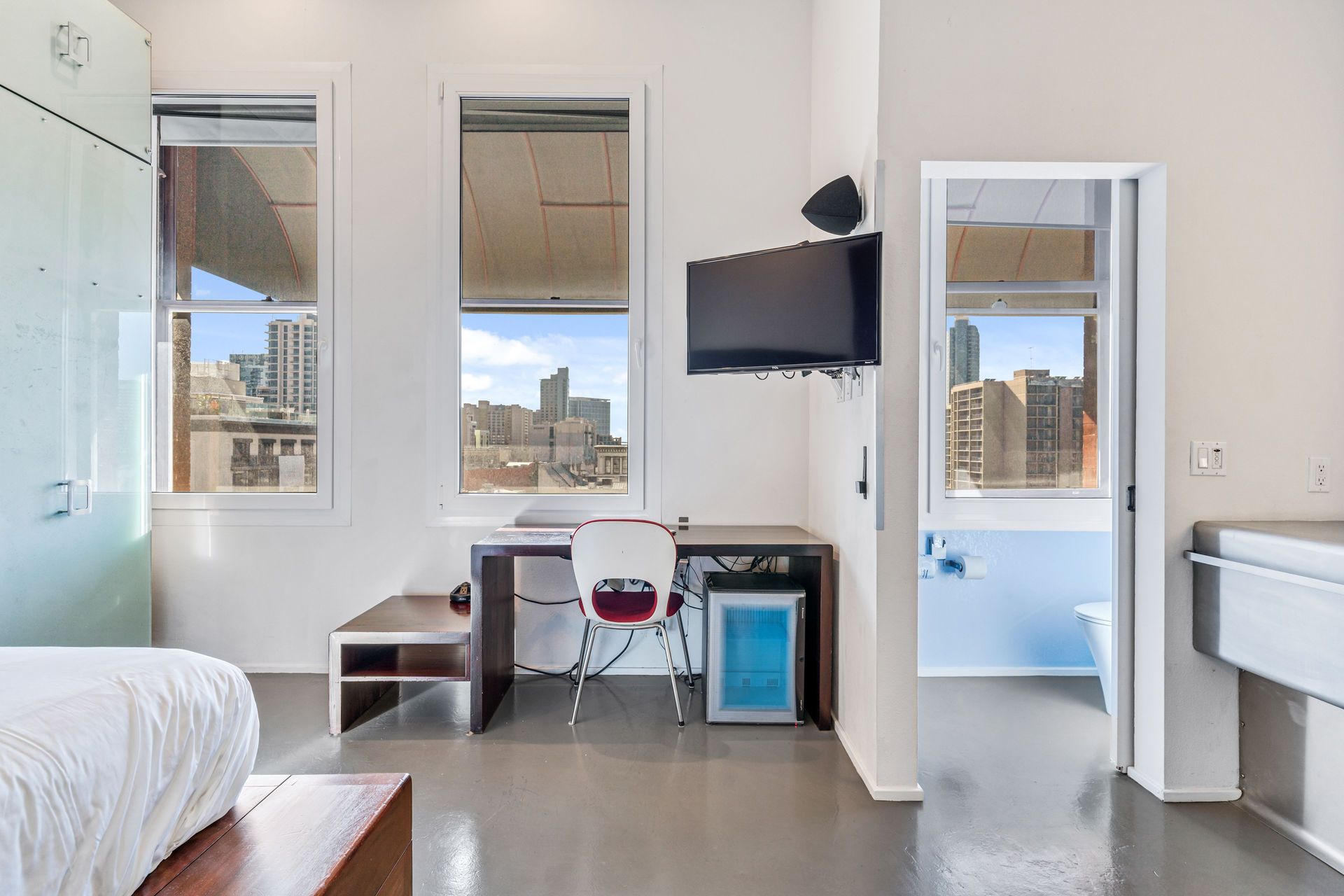 Bright room with a desk, chair, and mini-fridge near windows with a city view. A TV is mounted on the wall.