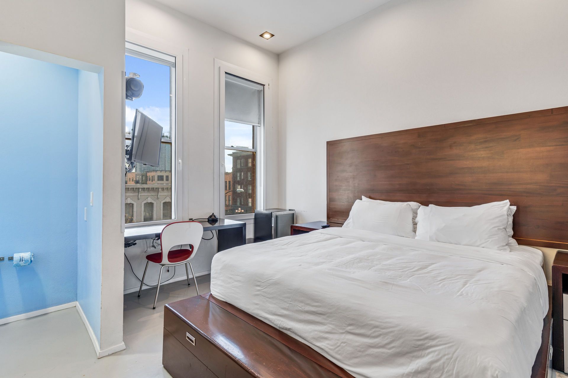 Bedroom with large bed, wooden headboard, small desk by window, bright white walls, and blue accent wall.